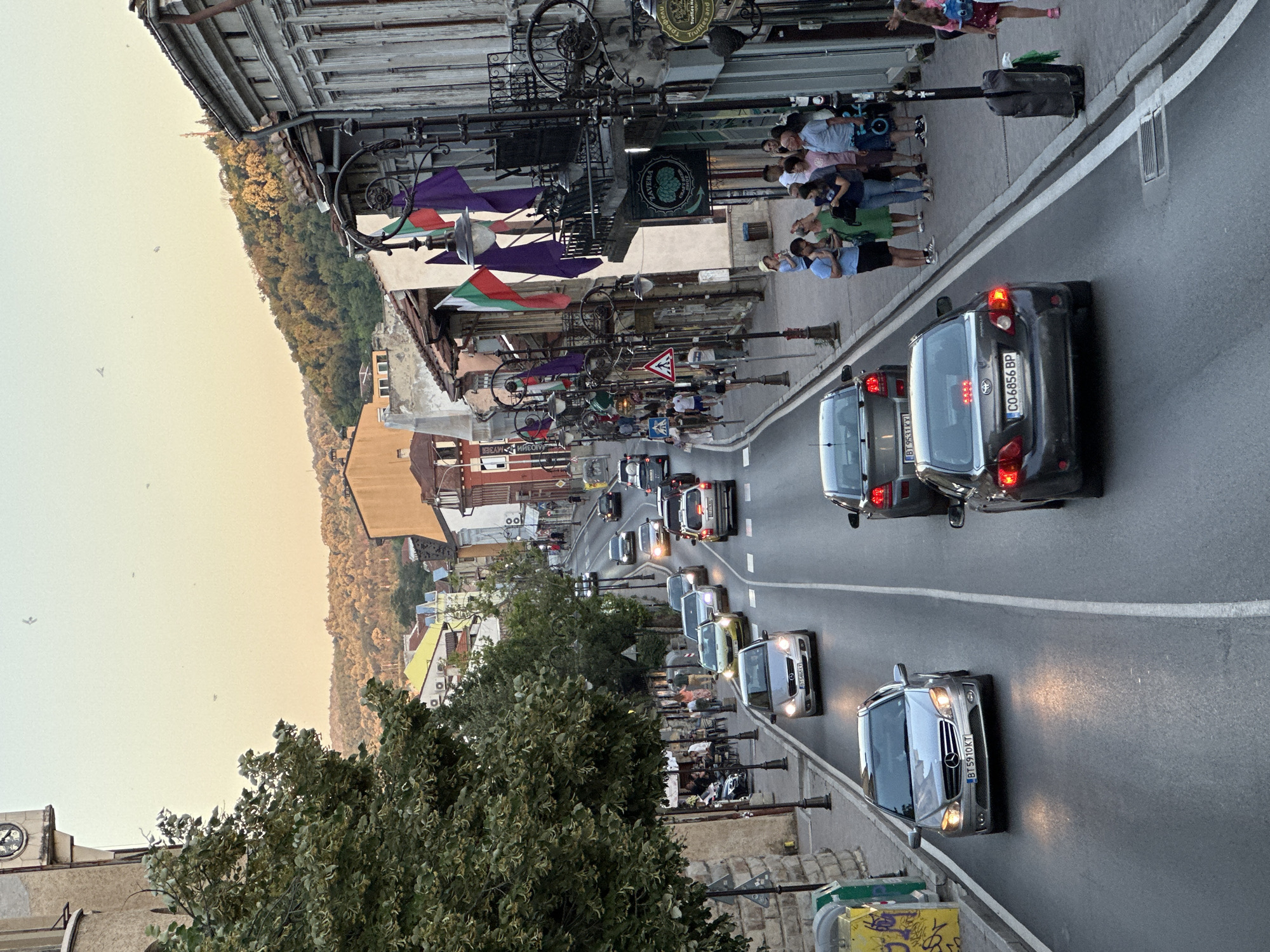 The image depicts a street scene in Veliko Tarnovo, Bulgaria, looking down a sloping road lined with buildings and trees. The perspective is from a higher vantage point, possibly a balcony or an elevated viewpoint. The street is busy with traffic, with several cars moving along the asphalt road. On the right side of the street, a sidewalk is visible with a group of people standing and walking, some of whom are looking at shops. The buildings lining the street have a historical architectural style, with some featuring ornate balconies and decorative elements. Bulgarian flags are prominently displayed on some of the buildings. The sky is a soft, warm hue, suggesting it might be late afternoon or early evening. The general atmosphere is one of a lively urban environment with a blend of modern and historic elements.