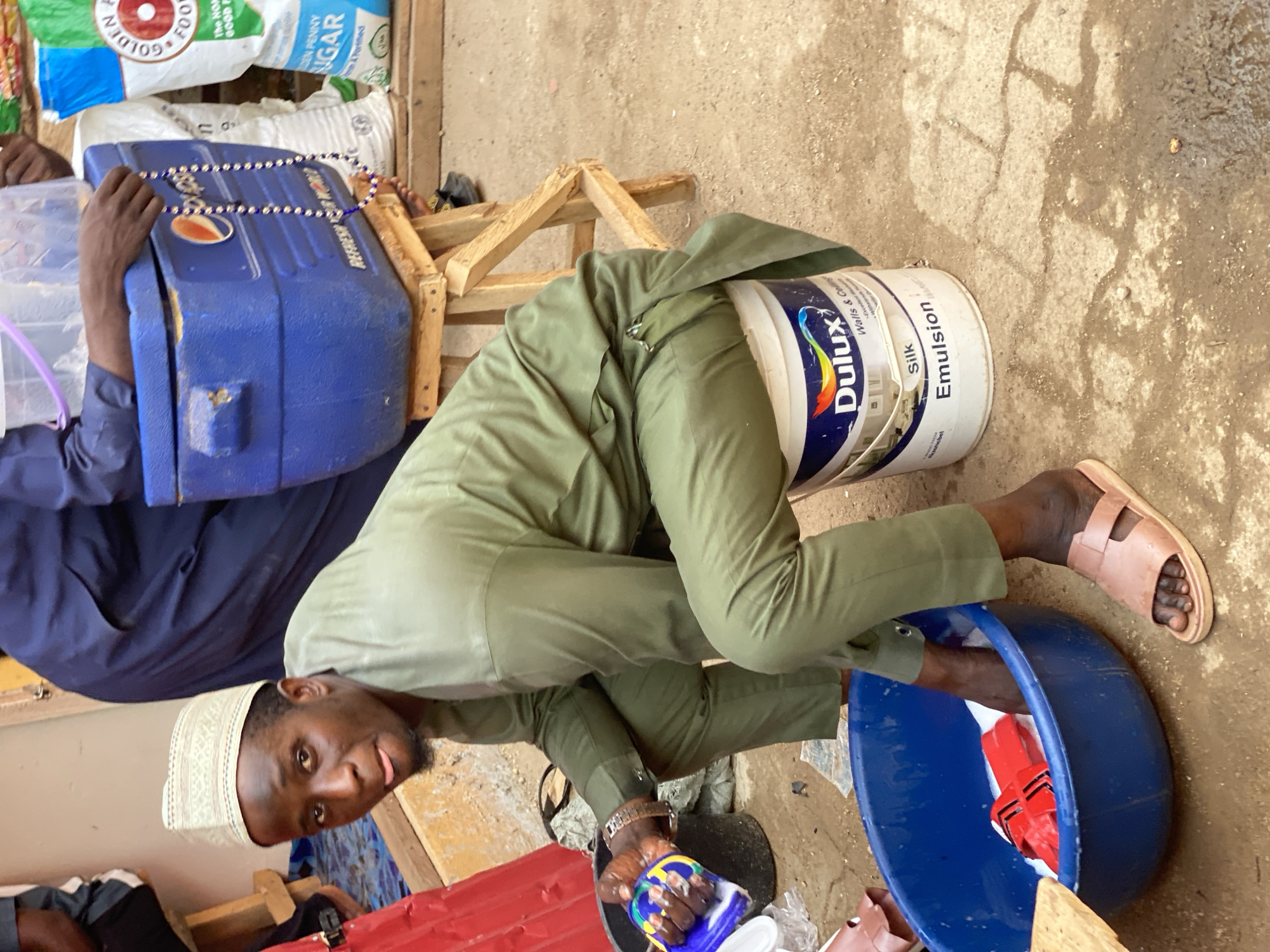 This image captures a vibrant market scene in Kafanchan B, Nigeria, showing a man engaged in an activity within an informal outdoor setting. The photograph is oriented sideways, giving an overhead perspective of the subjects and their surroundings.
The central subject is a man, likely in his 20s or 30s, dressed in a light green tunic and trousers, and wearing a white kufi cap. He is crouched down, looking directly at the camera with a neutral to slightly curious expression. His hands are positioned over a blue plastic basin, where a red object, possibly a tool or clamp, is visible amidst some liquid or suds. He holds a small blue and yellow container, partially identifiable by "Tide" branding, in his left hand. His right foot, wearing a light brown sandal, is visible next to the basin.
Beside him, a white Dulux paint bucket is prominent, bearing the text "Dulux Silk Emulsion Walls & Ceilings AkzoNobel." In the background, to the left, another person's arms in dark blue attire are visible, holding a large blue cooler adorned with a "Pepsi" logo and a string of blue and white beads, possibly a rosary. The background features various large sacks of goods, with visible text including "GOLDENA FOO," "The NOW GOOD E," and "DEN PENNY NGAR Ane Fortified." Wooden structures, possibly makeshift market stalls or furniture, are also scattered around. The ground is a mix of dirt and concrete, showing some damp patches. The scene suggests a busy marketplace or an informal trading area during daytime.