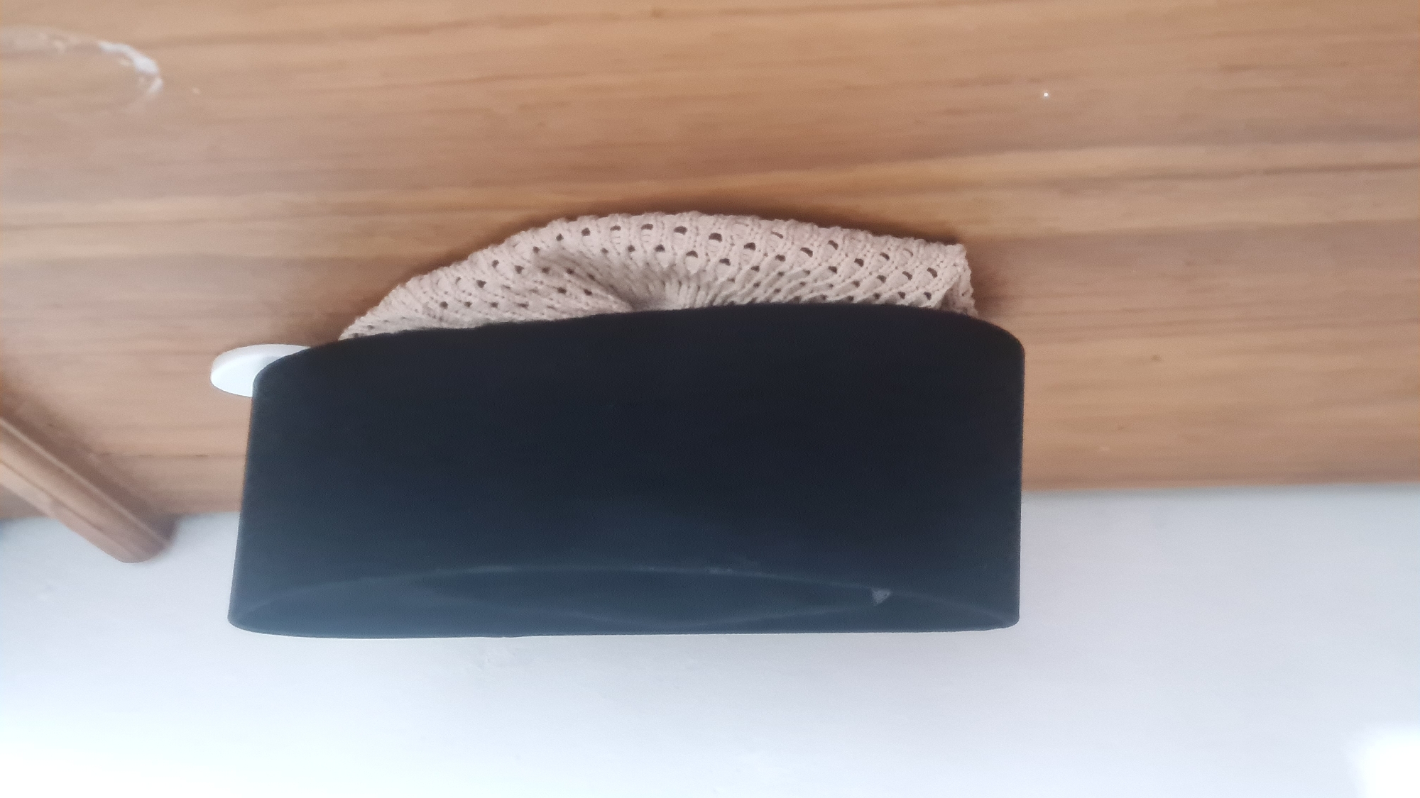 A black velvet hat with a white drawstring bag is hanging on a hook on a wooden surface, possibly a dresser or cabinet. The hat appears to be cylindrical with a flat top and a wide brim. The drawstring bag is made of a crocheted material and is loosely tied around the hat. The background is a plain white wall. The image appears to be taken indoors, and there is no indication of the location in Bandar Lampung, Indonesia.