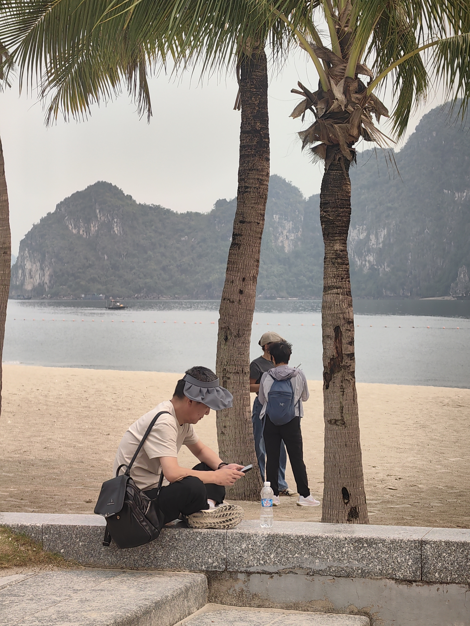 The image captures a serene coastal landscape in Ha Long Ward, Vietnam, featuring people relaxing amidst tropical scenery.
In the foreground, a man is seated on a granite ledge, his gaze directed downwards at his phone. He wears a light-colored short-sleeved shirt, black pants, and a grey sun visor. A black backpack is positioned next to him on the ledge, and a clear plastic bottle of "Aquafina" water is visible nearby. His right foot, clad in a patterned sneaker, rests on a woven straw object.
Behind him, on a light sandy beach, two other individuals stand between two large palm tree trunks. One person, seen from the back, wears a light jacket and a blue backpack, holding what appears to be a phone. They seem to be engaged in conversation with a partially obscured person standing closer to the tree, wearing a hat and a darker shirt.
The background features a calm, grey-blue bay dotted with a line of orange buoys and a small boat further out. Majestic limestone karst mountains, covered in greenery, rise from the water under a hazy, overcast sky, giving the scene a soft, diffused light. The prominent palm trees, with their textured trunks and green fronds, frame the composition. The overall mood is casual and relaxed.