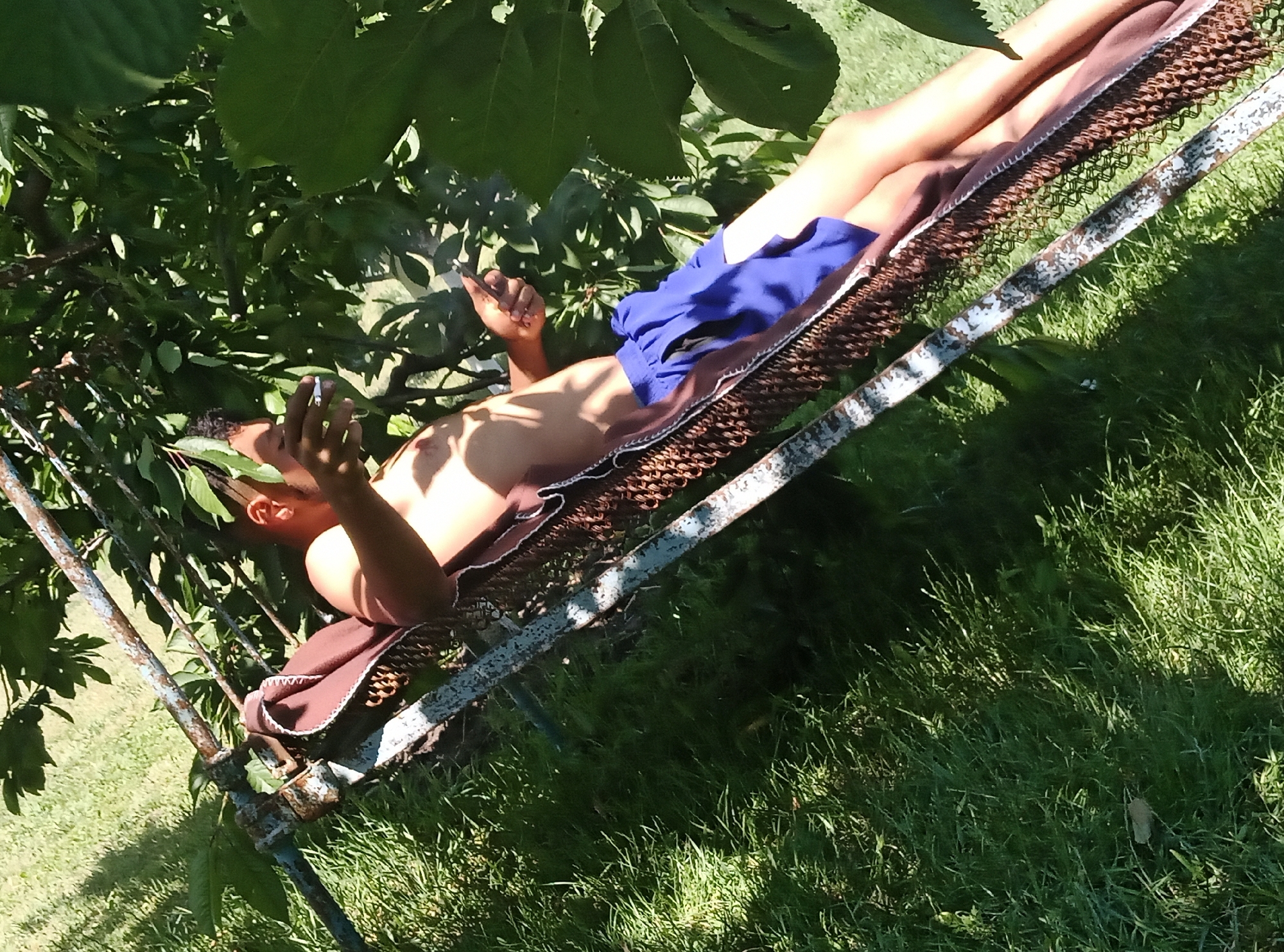 A person is lying on a hammock under trees in Cimișeni, Moldova. The person is on their back with their legs extended. They are wearing blue shorts and have a towel draped over their body. Their right arm is bent at the elbow and resting on their chest, while their left arm is extended. The person is holding a cigarette in their left hand, which appears to be lit. The hammock is made of a woven material and is supported by a metal frame. The background consists of green grass and leaves from the surrounding trees, suggesting an outdoor setting. The lighting indicates it is daytime, with sunlight filtering through the leaves, creating shadows.