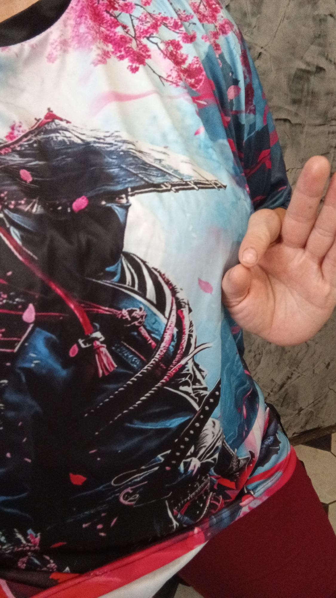 A close-up shot shows a person wearing a black, blue, and pink t-shirt with a samurai warrior print. The t-shirt is adorned with imagery of cherry blossoms and a mountain, possibly Mount Fuji. The person's right hand is visible, with the thumb and index finger forming a circle, while the middle, ring, and pinky fingers are extended. The background is a textured gray wall and a tiled floor. The image was taken in Drezna, Russia.