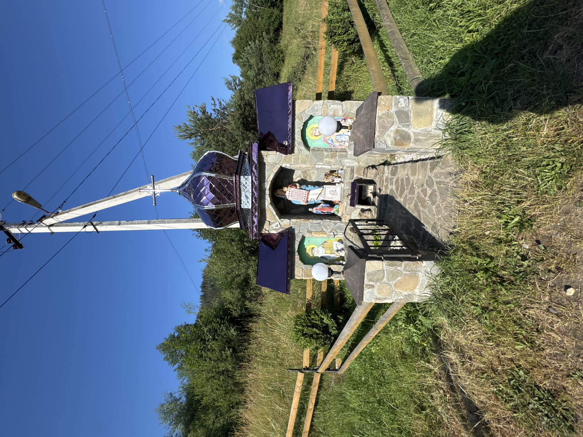 A roadside chapel, likely a votive shrine, stands under a clear blue sky in Kryvorivnia, Ukraine. The structure is built of rough-hewn stone and features a small, arched entryway housing religious icons and figurines. A purple, faceted dome crowns the chapel, and a cross is visible on top. Two white, spherical lamps are mounted on stone pillars flanking the entrance. A wooden fence runs behind the shrine, and the surrounding area is overgrown with grass and weeds, with trees and a hillside visible in the background. A power pole with visible wires stands to the right of the chapel, casting a shadow that partially obscures the foreground. The image appears to have been taken on a sunny day.