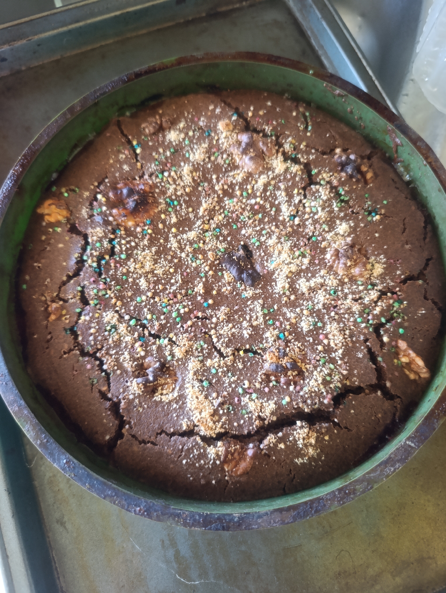 A close-up, overhead shot shows a round, dark chocolate cake with walnuts and colorful sprinkles baked in a round, dark green cake pan. The cake has a cracked surface, and some of the sprinkles appear to be dissolved into the cake. The cake pan is set on a metallic surface, possibly a baking sheet. The background is blurred, but suggests an indoor setting. The image was taken in Olomouc, Czechia.
