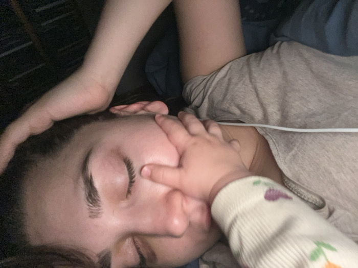 An overhead close-up captures an adult individual, eyes closed, lying supine. The individual wears a light brown t-shirt. Their left arm is bent, with the hand positioned near the top of the head. A small child's left hand is placed over the adult's left eye and upper cheek, with fingers pressing into the skin. The child's forearm is partially visible, clothed in a white garment with faint multi-colored patterns, including purple and orange elements. A white cable rests across the adult's upper chest, near the collarbone. The immediate environment includes blue fabric, likely bedding, and a dark, indistinct background. The scene is dimly lit.