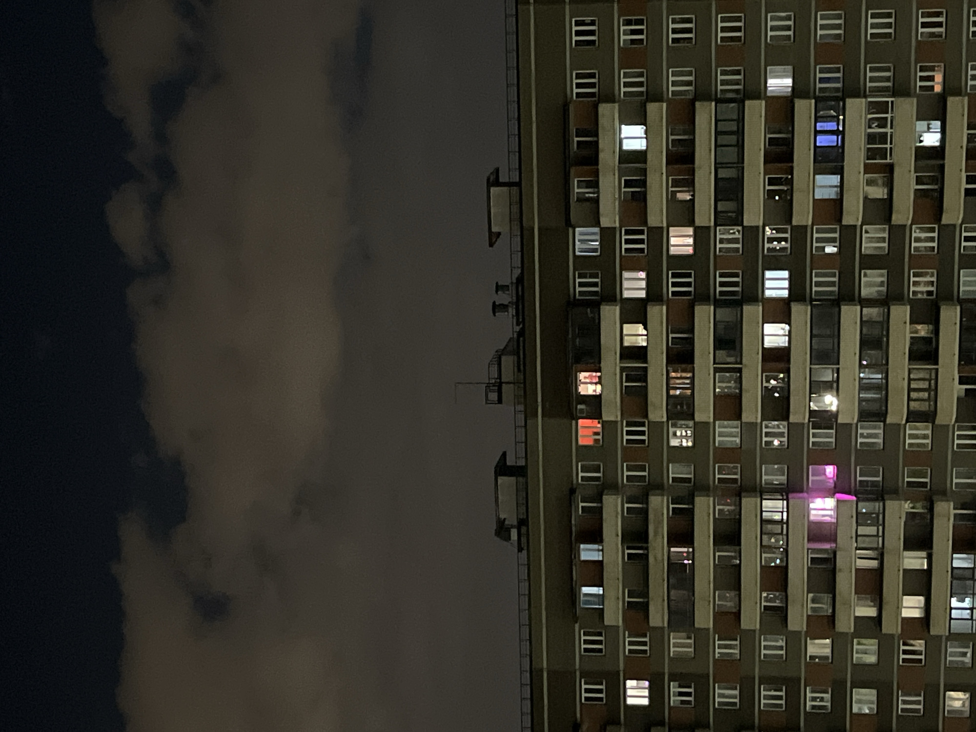 The image presents a high-rise building at night, contrasted against a dark sky and clouds. The building is oriented vertically in the right half of the frame, while the left half is dominated by a dark sky with cloud cover. The building is multi-storied, with numerous windows visible, some illuminated from within with varying colors like white, blue, red, and pink. The architecture appears utilitarian and repetitive. A fire escape is visible on the side of the building. The location is likely urban.