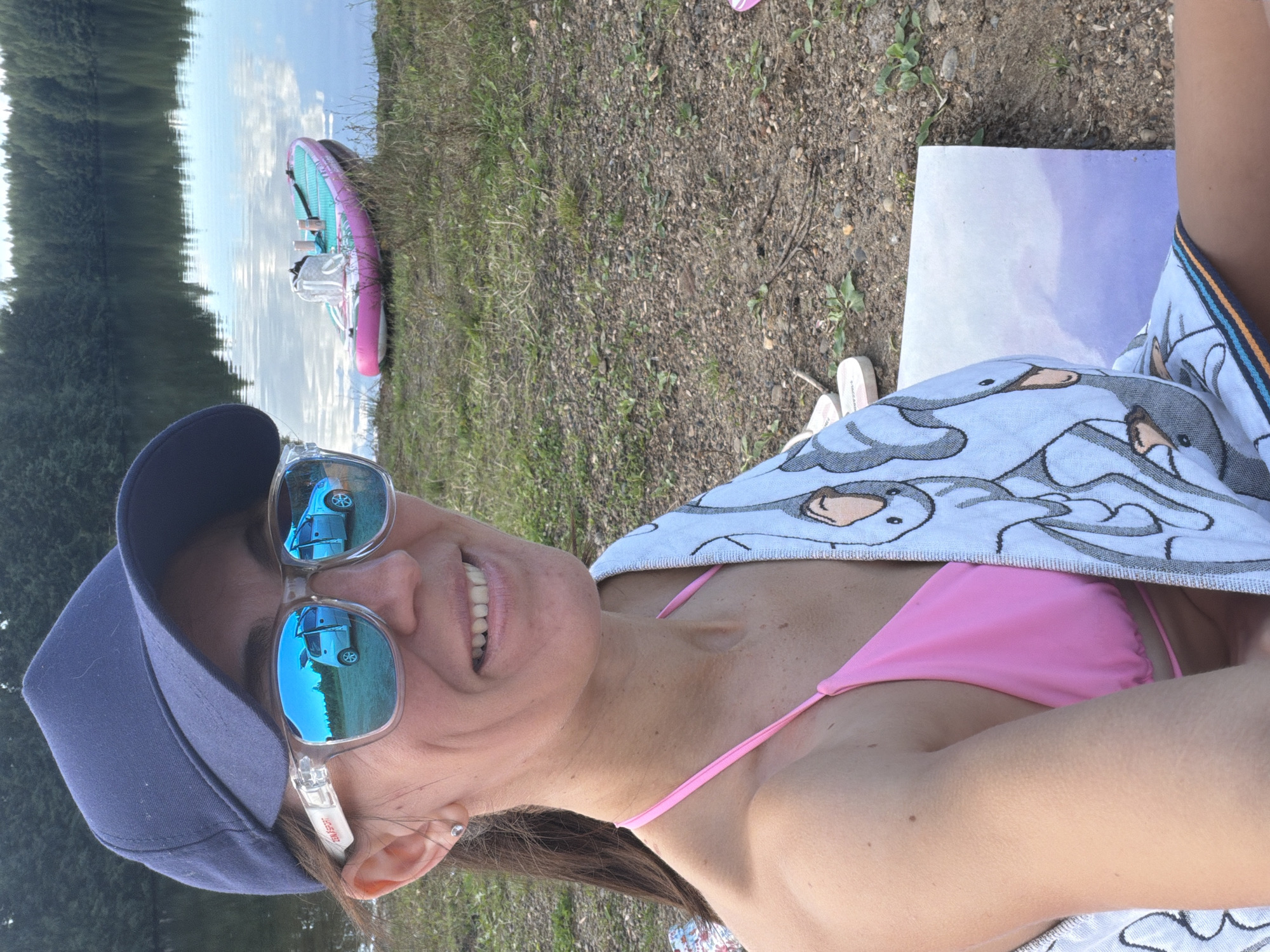 In Chernushka, Russia, a woman with a dark blue baseball cap and clear-framed sunglasses smiles at the camera. Her sunglasses reflect a silver car with its doors open, parked on a grassy area near a calm body of water. She is wearing a pink bikini top and has a towel with a cartoon goose print draped over her shoulder. In the background, a pink and turquoise paddleboard rests on the shore of the lake, with trees lining the opposite bank. The ground is covered in dirt, pebbles, and sparse green vegetation.