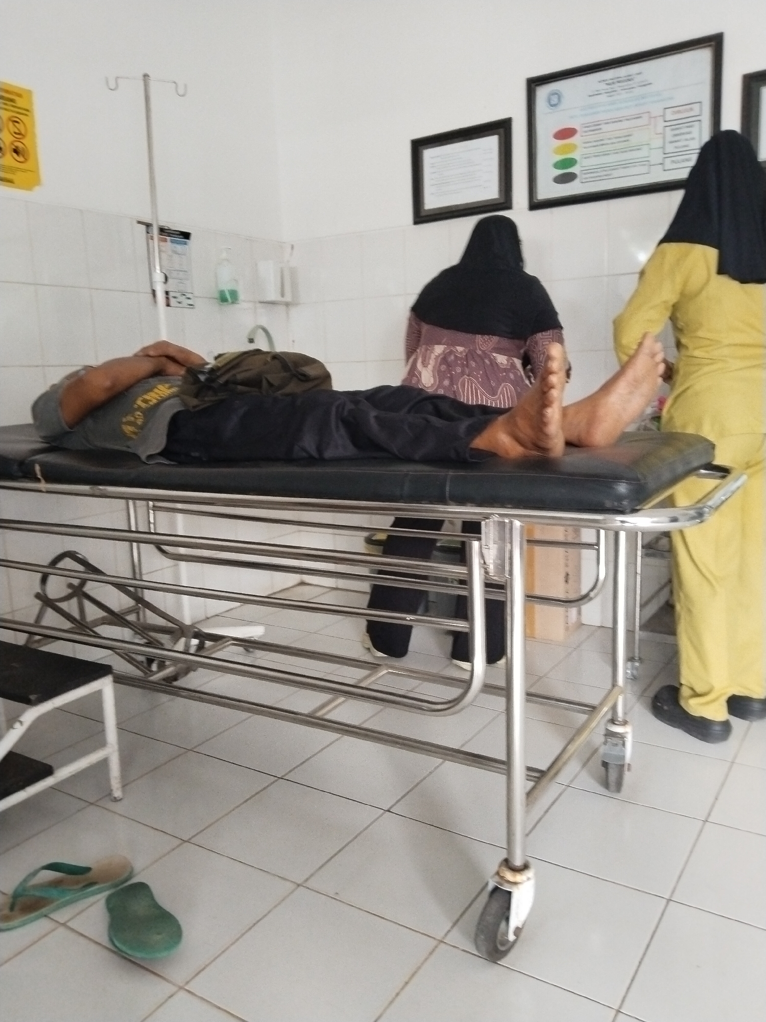 The image shows a patient lying on a stainless steel gurney in what appears to be a medical facility, possibly in Prigi, Indonesia. The patient is a male, lying on his back with his hands crossed over his chest. He is wearing a gray t-shirt and dark pants. A green bag is placed on the gurney next to him. Two women are standing in the background, both wearing hijabs. One woman is wearing a patterned top and a dark hijab, and the other is wearing a yellow uniform and a black hijab. The room has white tiled walls and floors. There is an IV stand to the left of the gurney, and a framed poster on the wall. In the foreground, a pair of blue and green flip-flops are on the floor. The overall scene suggests a medical setting where a patient is receiving care.