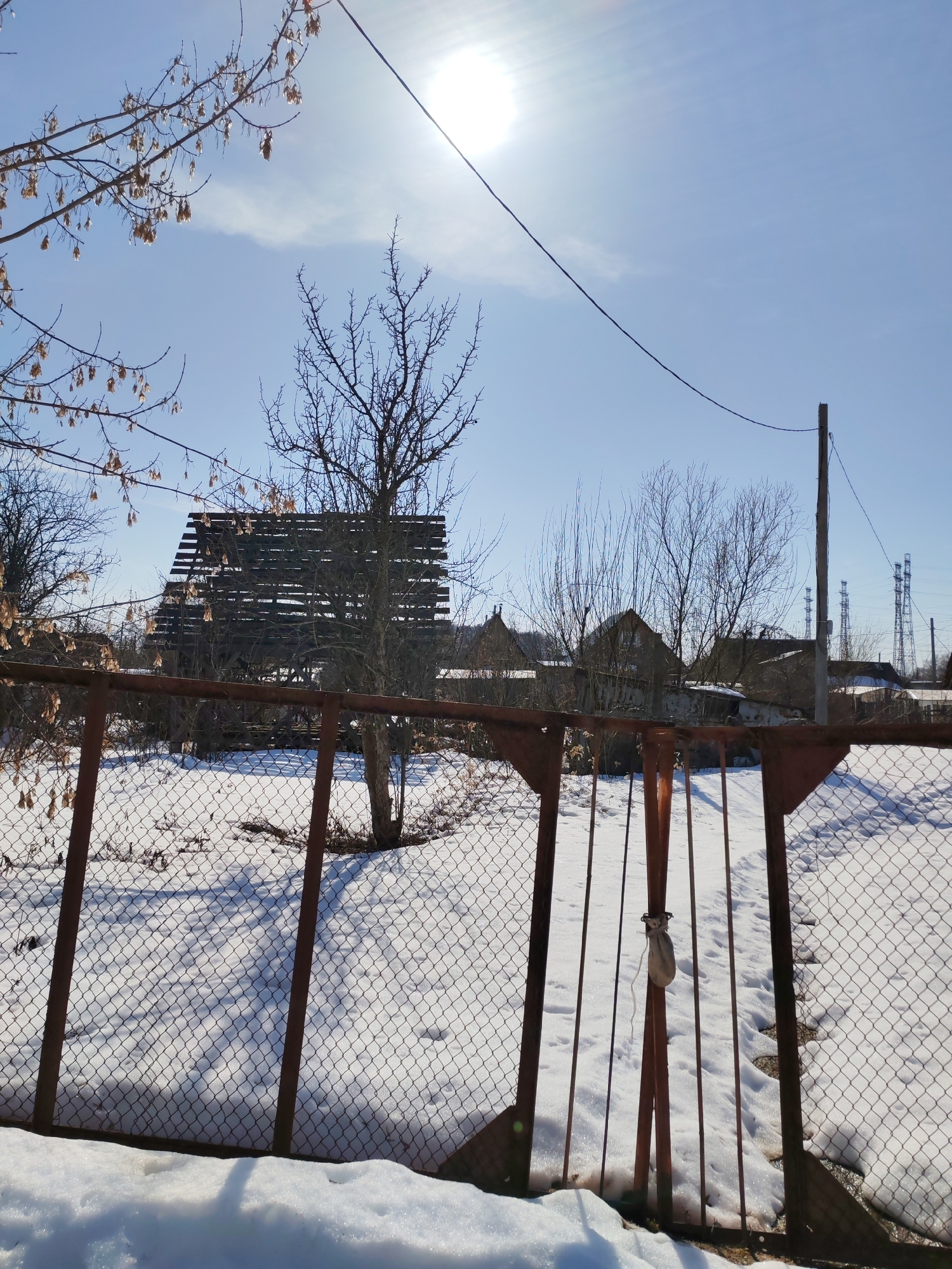 The image depicts a winter scene in what appears to be a residential area. A rusty, metal chain-link fence dominates the foreground, with a closed gate secured by a small bag. Behind the fence, the ground is covered in snow. Several bare trees and shrubs are scattered throughout the scene. A wooden structure with slats is visible behind the trees, partially obscured by branches. In the background, houses and utility poles can be seen. The sky is bright blue with the sun shining. The location is Chaikovsky, Russia.