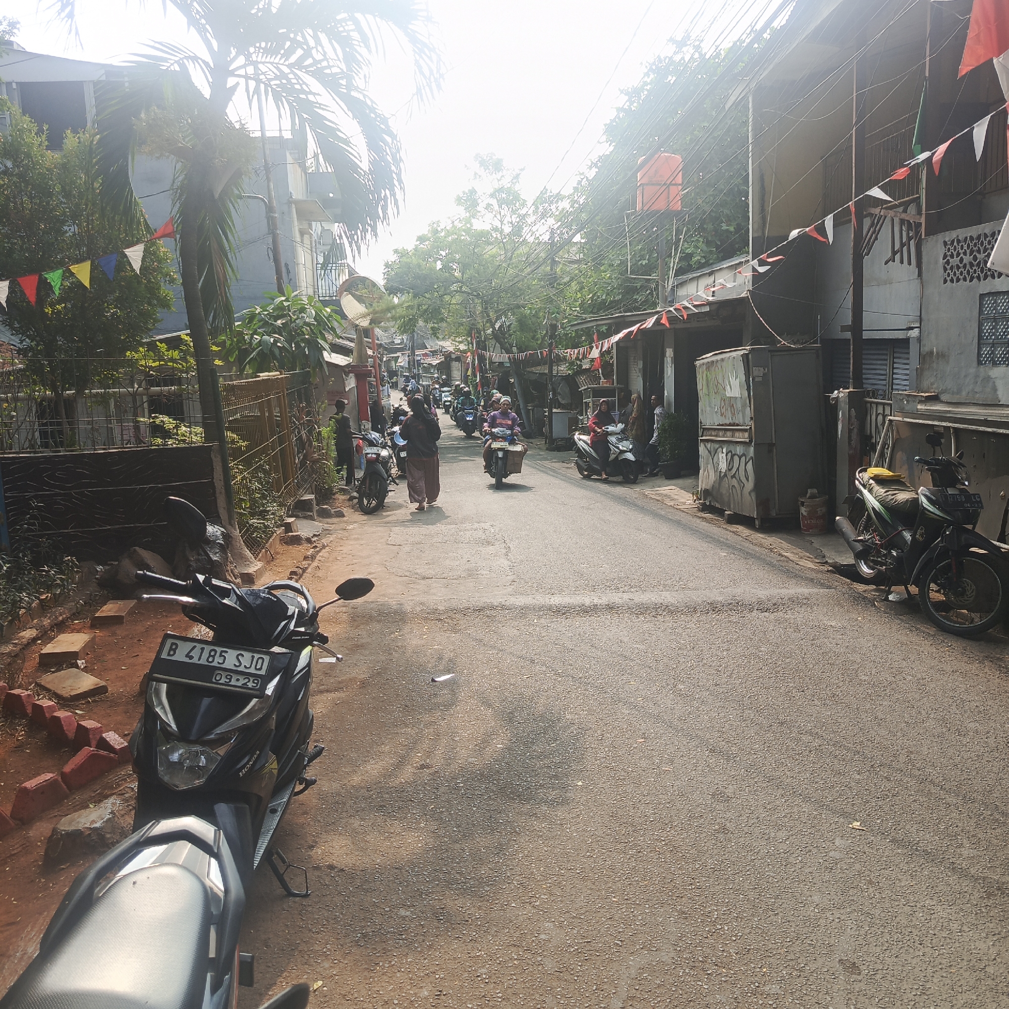 The image depicts a narrow street in a residential area of South Jakarta, Indonesia, likely during daytime given the bright sunlight and shadows. The street is paved with asphalt and lined with modest buildings, many of which appear to be two stories high, with visible electrical wires and conduits overhead. Decorative flags, predominantly red and white, are strung across the street and along the building facades, suggesting a festive occasion or national celebration.
Several motorcycles are present, both parked and in motion. In the foreground, a black Honda scooter with license plate "B 4185 SJO" is parked on the side of the road. Further down the street, other motorcycles are seen with riders, indicating normal daily activity. People are visible walking and riding motorcycles, dressed in casual attire, with some women wearing headscarves. One individual is seen pushing a motorcycle.
The environment is a mix of urban and natural elements, with some trees and vegetation visible, including a palm tree on the left. A large orange water tank is mounted on a pole on the right side of the street. Buildings show signs of wear and tear, with some featuring decorative metalwork or patterned walls. A convex mirror is mounted on a pole near the left side of the image, likely to improve visibility at an intersection. A temporary stall or kiosk is also present on the left side of the street. The overall impression is of a busy, lived-in urban environment characterized by its dense population and active street life.