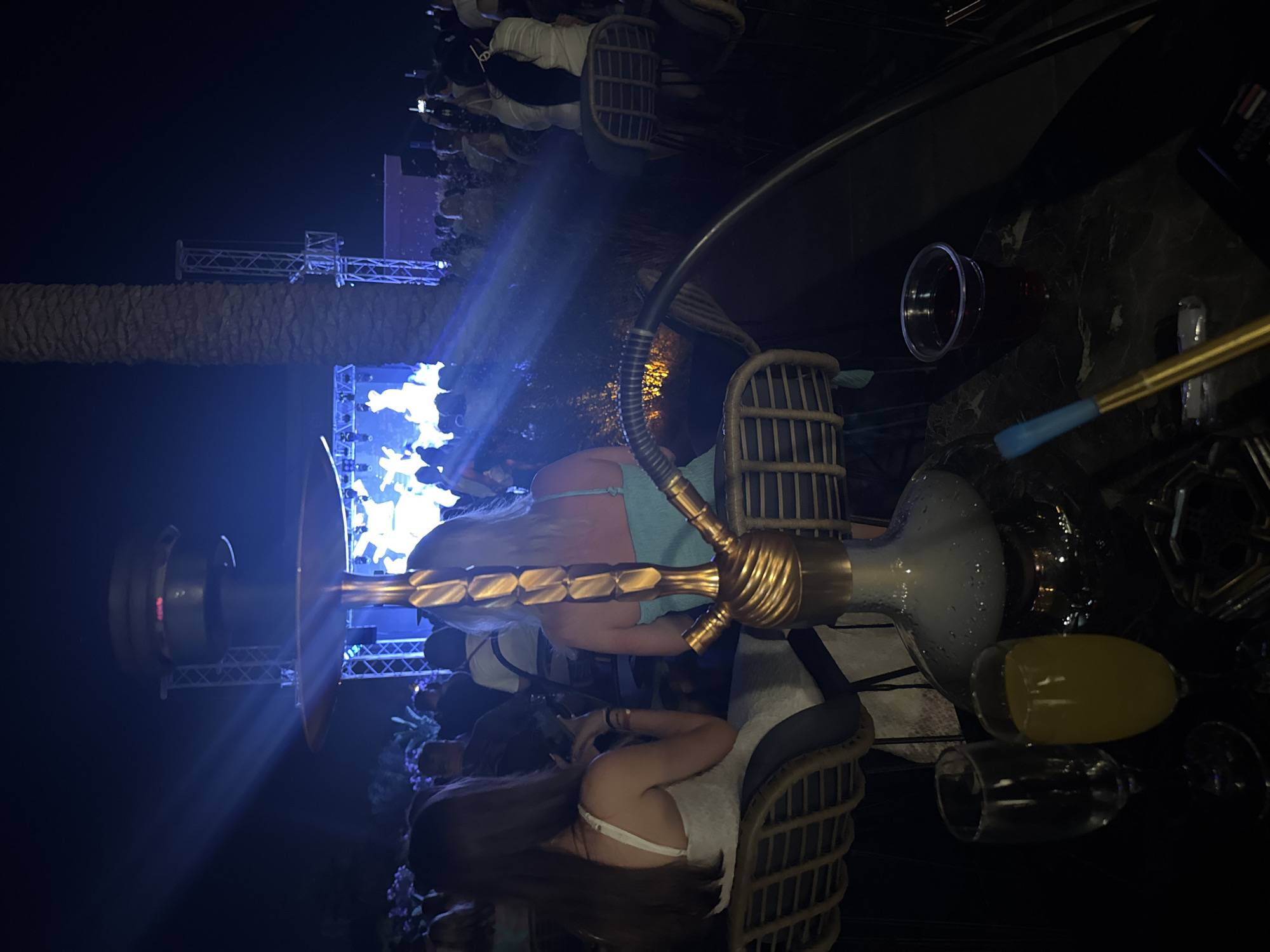 A wide shot captures a nighttime outdoor concert scene in Kemer, Turkey, viewed from a table with a hookah pipe and drinks. In the foreground, the ornate, gold-colored hookah pipe is prominently featured, positioned on a dark, reflective surface with a glass of amber liquid, an empty champagne flute, and a small ashtray with what appears to be a blue-tipped hookah hose. Two women are seated at rattan chairs, facing away from the camera, with their backs to the viewer. One woman on the left wears a white dress, and the woman on the right wears a turquoise top. Behind them, a large crowd is gathered, facing a brightly lit stage where a performance is underway, indicated by the illuminated figures on stage and the intense blue stage lighting that casts beams into the dark sky. The stage is flanked by industrial-looking metal structures. A large palm tree trunk is visible to the right of the stage, adding to the tropical ambiance of the location. The overall atmosphere suggests a lively entertainment venue.