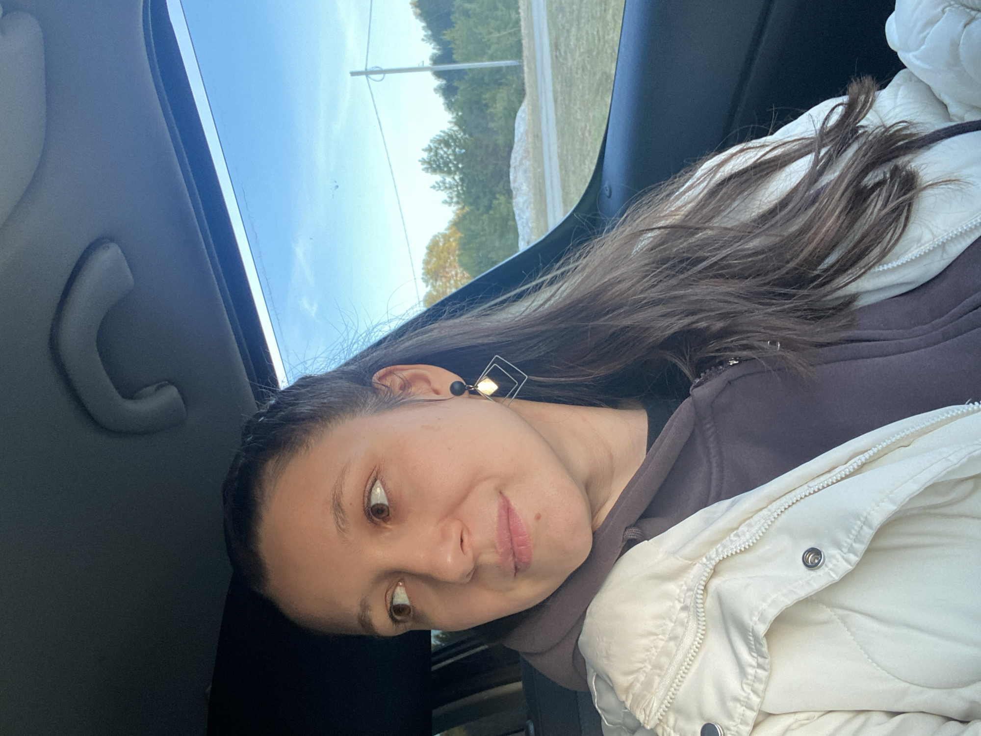 The image depicts a young woman inside a car, captured in Chaikovsky, Russia. The photograph is presented rotated 90 degrees clockwise from its upright orientation. The woman is the primary subject, occupying the right side of the frame (when viewed rotated). She has long, dark brown hair cascading over her shoulders and part of her attire. She is dressed in a dark brown hooded sweatshirt and an off-white, quilted puffer jacket with a zipper and snaps. A distinctive earring, featuring a black stud and a silver-toned square outline, is visible on her right ear.
Her face is turned slightly to her left, with her brown eyes looking directly forward, exhibiting a subtle, contemplative expression with a slight upturn at the corner of her lips. The car's interior, showing a light grey ceiling and a grab handle, is visible on the far left.
Through the car window, an outdoor scene unfolds under a bright blue, clear sky. A roadside landscape is visible, featuring green and yellowish-green trees, a patch of light-colored ground or dry grass, and a paved road in the distance. A utility pole with wires is also discernible against the sky, indicating a rural or suburban roadside setting. The sunlight suggests it is daytime, possibly late morning or afternoon. No text is visible in the image.