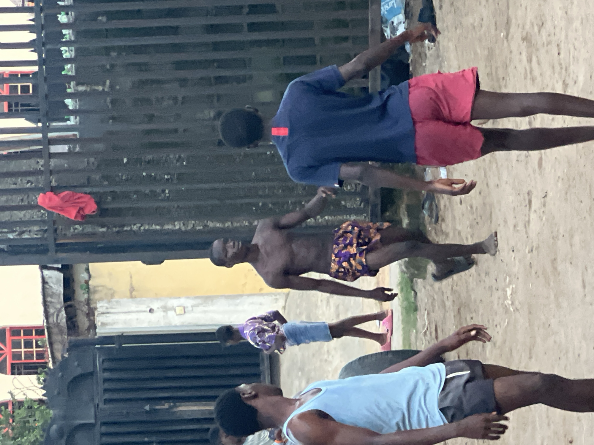 The scene is outdoors in Ijegun, Nigeria. Four Black males are visible, seemingly engaged in physical activity. The first, wearing a blue shirt and red shorts, is at the top of the image. The second male is shirtless, wearing patterned shorts. The third male wears a patterned shirt and blue shorts with pink sandals. The fourth male wears a blue tank top and dark shorts, positioned near a tire. A metal gate is in the background, along with a building with red window frames. A red cloth hangs from the gate. The ground is sandy or dirt-covered.