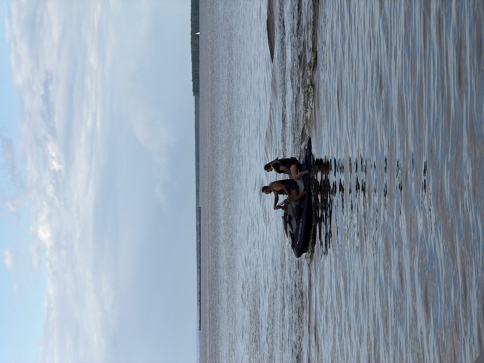 The image captures a serene daytime scene on a large body of water, likely the Kama River in Chaikovsky, Russia, given the context. Two individuals, a man and a woman, are riding a jet ski. The man, wearing sunglasses and a dark vest, is at the helm, while the woman, also in a vest, is seated behind him. They are both facing forward, navigating the choppy water. The jet ski is a dark color with the registration number "AA5128RUS59" visible on its side. In the background, a long barge is visible in the distance, stretching across the water. Further still, a treeline marks the far shore of the river, with a small sailboat adding a touch of distant activity. The sky above is partly cloudy, with soft white clouds scattered across a pale blue expanse. The water itself reflects the sky and surrounding environment, creating a textured, rippling surface.