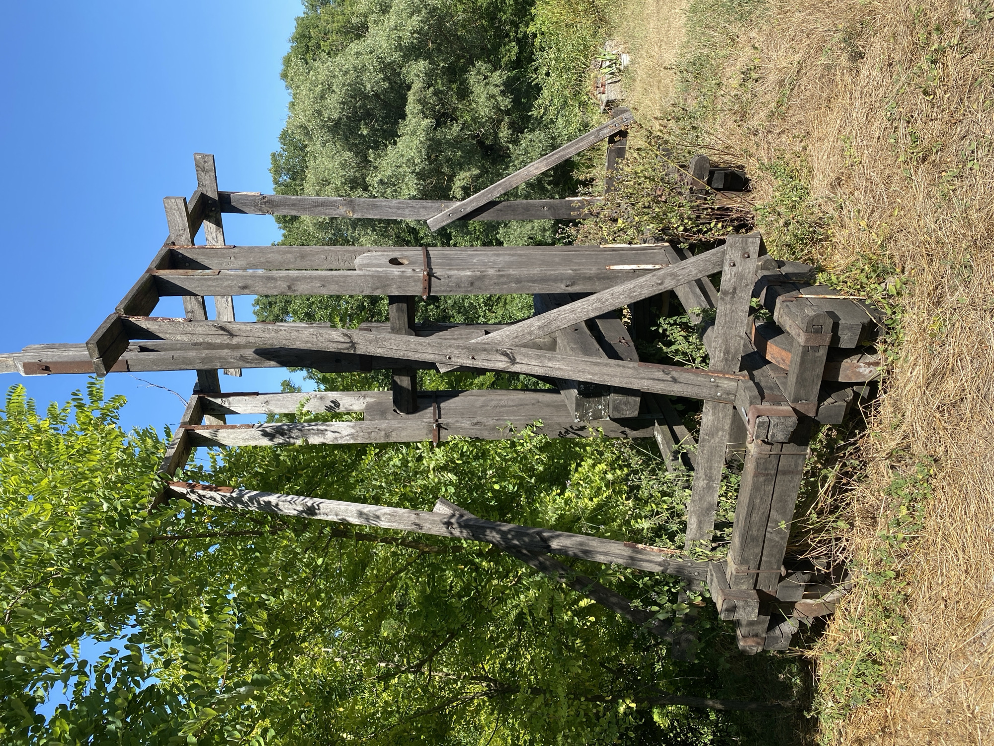The image depicts a weathered, wooden structure resembling a trebuchet or catapult, located outdoors in what appears to be a rural or overgrown area. The structure is constructed from dark, aged wood and metal hardware, showing signs of disrepair and exposure to the elements. It features a large, upright frame with various beams and supports, and a wheeled base that suggests it might be mobile.
The surrounding environment consists of dry, yellowish grass and dense green foliage from trees and bushes. The sky is a clear, bright blue. In the background, more trees and vegetation can be seen, creating a natural, somewhat wild setting. There are no people or animals visible in the frame. The overall impression is one of an abandoned historical artifact or a prop set in a natural landscape.
The location is identified as the city of Busha, Ukraine, which is known for its historical and cultural sites. This particular structure is likely a relic or a reconstruction related to the region's past, possibly a historical reenactment site or a monument. The image captures the object in daylight, under natural lighting conditions.