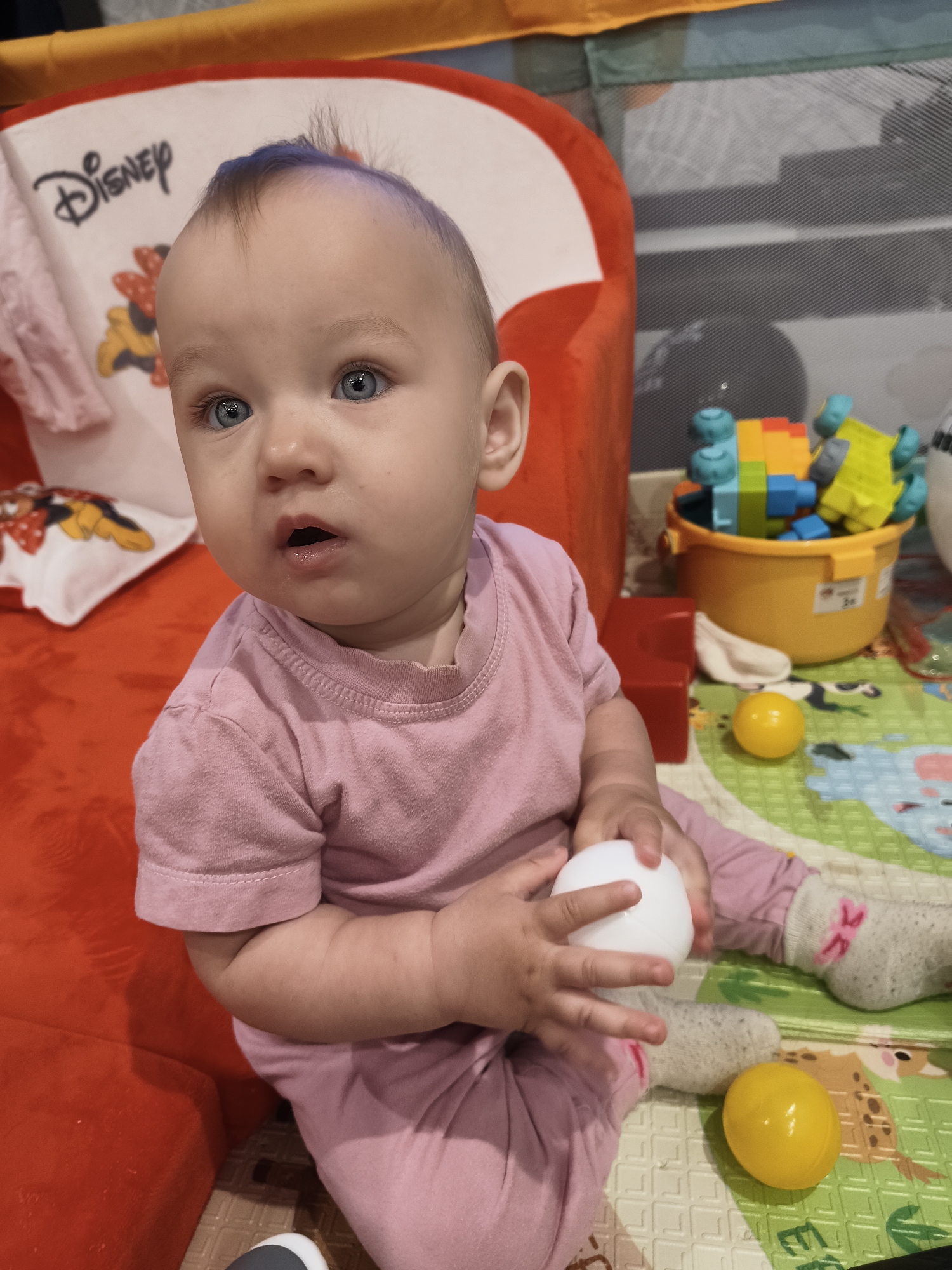 A close-up, eye-level shot shows a baby with blue eyes and light brown hair, some of which appears to be dyed blue, sitting on a red surface, possibly a couch or playmat. The baby is wearing a pink t-shirt and pink pants, with gray socks that have a pink bow pattern. The baby is holding a white ball in its hands. The background includes a playpen with colorful toys, including building blocks and a yellow bucket. A mat with animal illustrations is visible on the floor. The scene appears to be indoors, likely in a home environment. The location is noted as Krasnogorskoye, Russia.