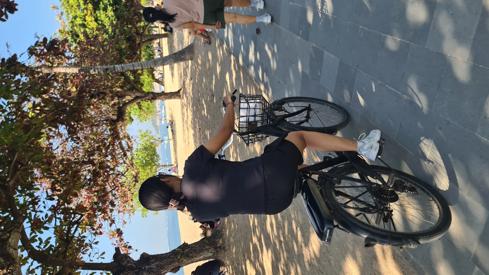 An outdoor, eye-level, medium shot shows a person riding a bicycle on a paved path next to a sandy beach. The rider, seen from the rear and right, wears a black t-shirt and black shorts. They have dark hair tied back with a light-colored scrunchie. White athletic shoes are visible on their feet. The bicycle is black with a front basket containing white items. The background features a sandy beach with scattered trees and the ocean. The sky is clear and blue. In the distance, on the beach, other individuals are visible, some sitting and some walking. Another person, also seen from the rear and right, walks on the paved path ahead of the cyclist, wearing a light brown t-shirt and olive green shorts with white socks and athletic shoes. The scene is lit by bright sunlight, creating shadows on the paved path. The location appears to be a coastal area in Denpasar, Indonesia, likely a popular spot for recreation.