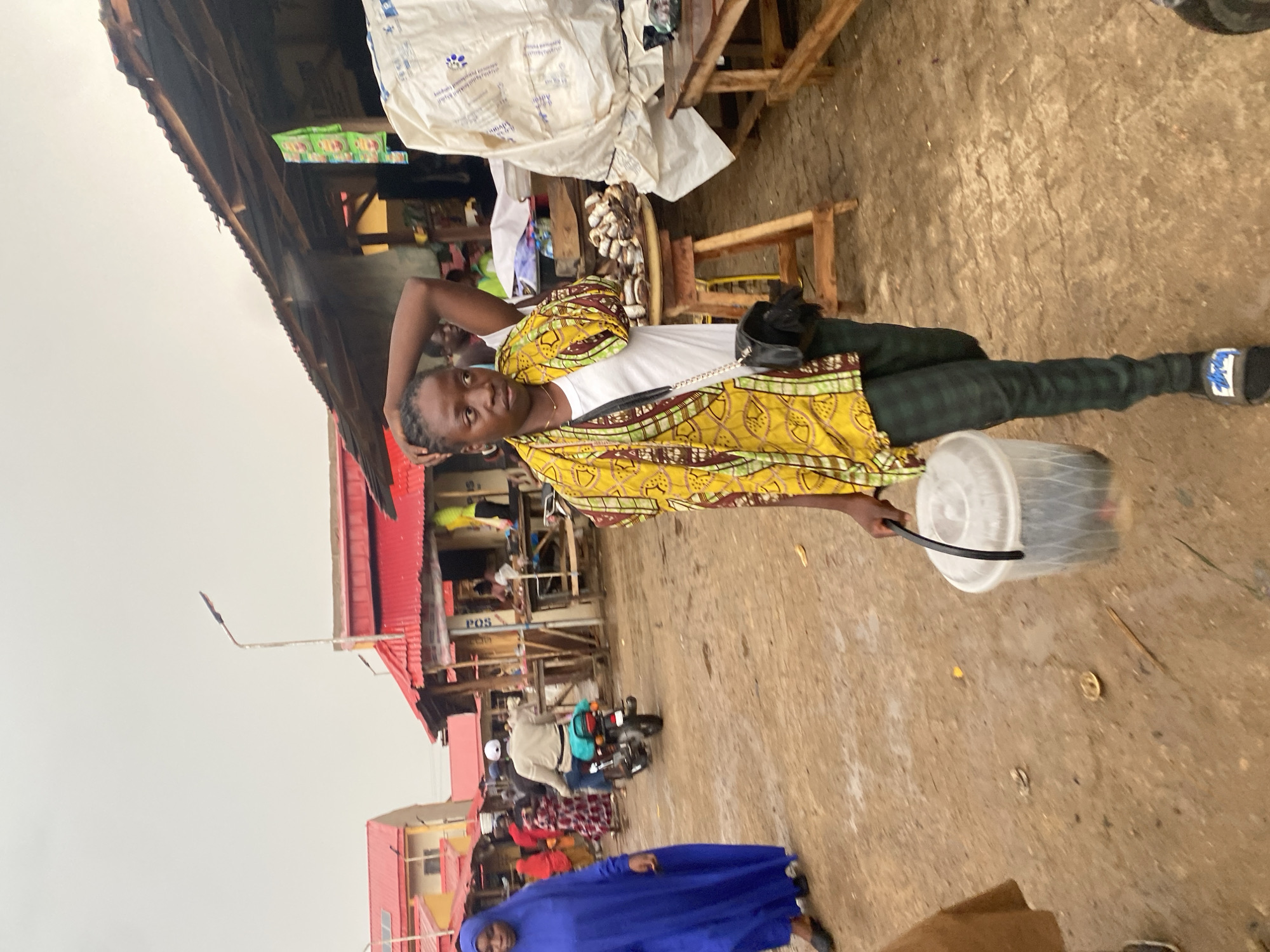 The image captures a scene in Kafanchan B, Nigeria. A woman stands in the foreground wearing a yellow patterned shirt, a white top, and checkered pants. She has a black purse slung across her shoulder and is holding a clear plastic container. The background reveals a bustling marketplace with stalls and people. There is a building with a red roof and "POS" written on it. A person on a motorcycle and a woman in a blue robe are also visible in the lower part of the frame. The ground appears to be paved or made of concrete. The weather seems overcast.