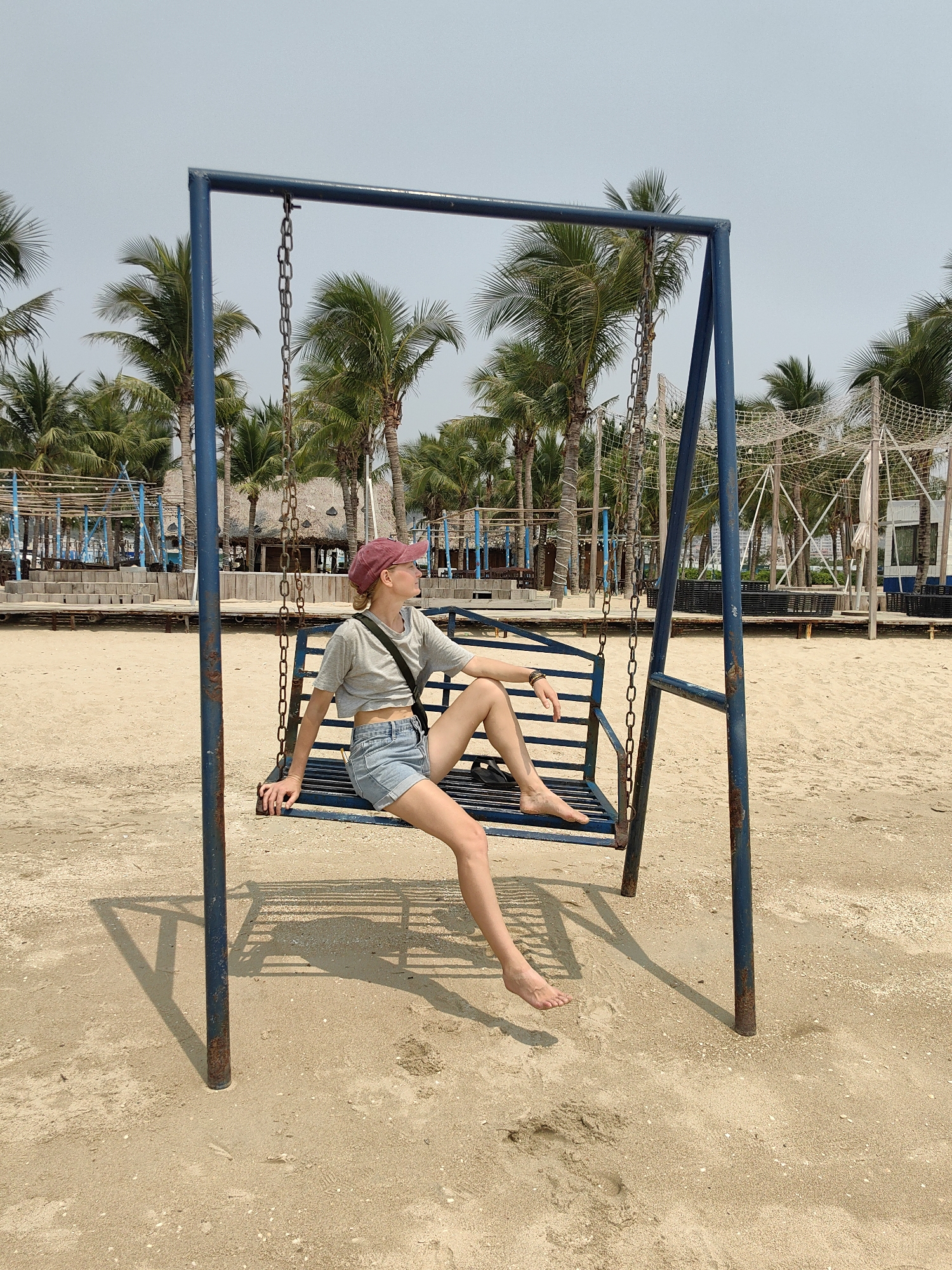 A full shot captures a fair-skinned woman with fair hair, adorned with a maroon baseball cap, sitting barefoot on a swing set on a beach. She is wearing a grey t-shirt, denim shorts, and a black crossbody bag. The swing set, made of blue metal, is on a sandy beach populated with palm trees and lounge areas with wooden structures. In the background, one can see the cityscape of Bai Chay Ward, Vietnam. The weather appears sunny, casting sharp shadows on the sand.