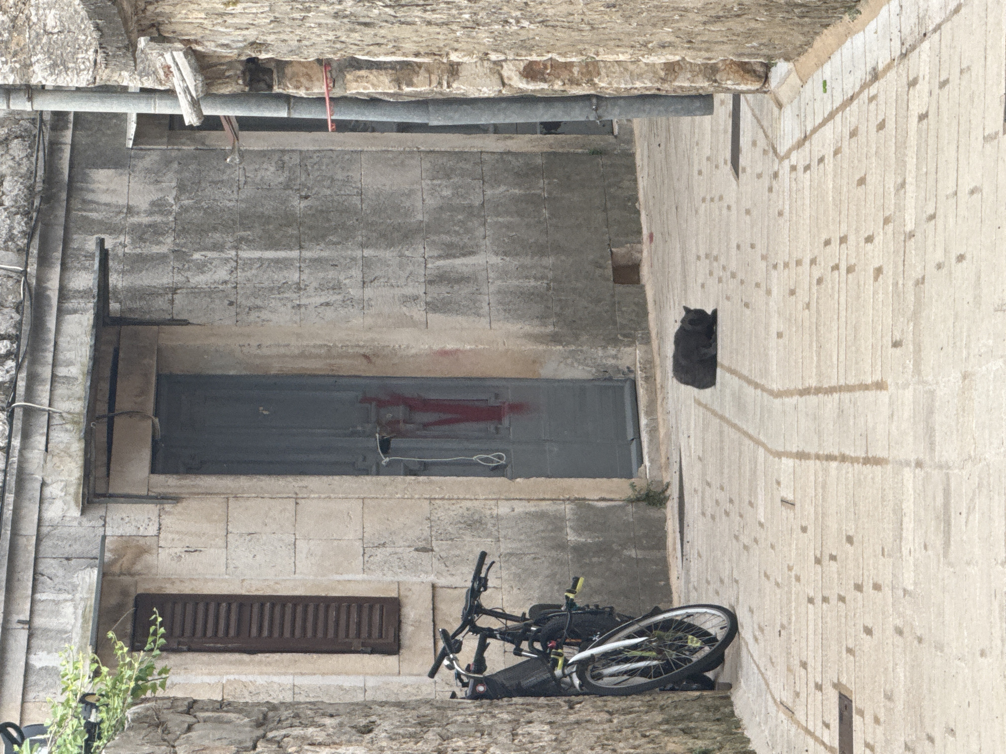In the city of Grad Stari Grad, Croatia, a narrow cobblestone alleyway is depicted. The scene is dominated by weathered stone buildings on either side. On the left, a stone wall partially obscures a building with a dark, louvered window. Leaning against the wall are two bicycles, one a darker color with a visible front suspension and the other a lighter color with a green rim.
The right side of the alley features a textured gray stone wall with a large, dark gray door that has a red "V" shape painted on it. A white rope is tied to the door handle. A vertical metal pipe runs alongside this building.
In the middle of the alley, a dark gray cat is resting on the cobblestones, facing left. The cobblestones are laid in a pattern of rectangular stones with smaller square stones interspersed. The lighting suggests an overcast day.