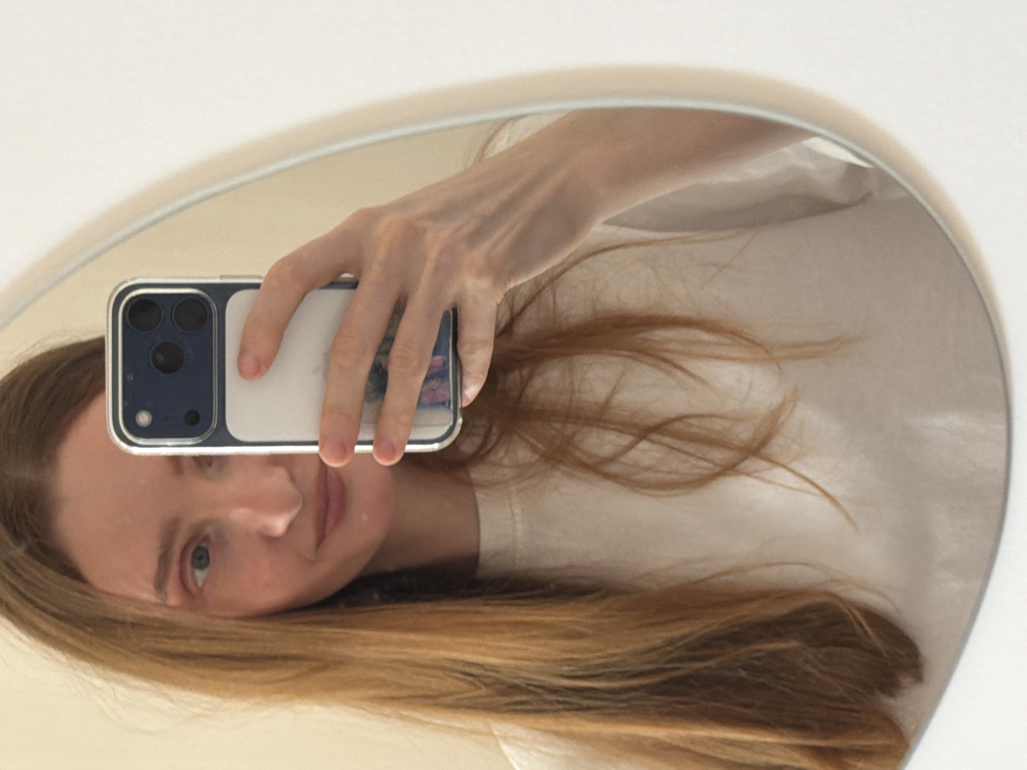 A woman with long, light brown hair is taking a selfie in a mirror. She is holding a phone with a clear case that has a blue edge. The phone has three cameras on the back. The woman is wearing a white t-shirt. The background is a white wall, the location is unknown but likely indoors. Boreti, Montenegro is not indicated in this image.