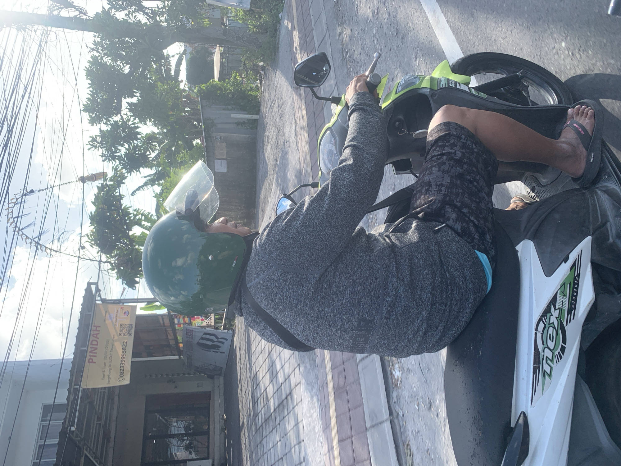 A person is riding a bright green and white scooter on a street in Denpasar, Indonesia. The rider is wearing a green helmet and a grey jacket. The scooter has a sticker that says "nex" in green lettering on the side. In the background, there is a building with a sign that says "PINDAH" in red letters. The sky is cloudy and there are trees on the right side of the image.
