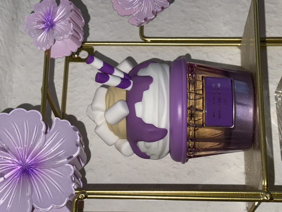 This image, located in Vellmar, Germany, depicts a decorative object resembling a dessert on a golden metallic shelf, against a textured, light-colored wall. The primary subject is a purple and white dessert-themed ornament, appearing to be a sundae or milkshake. It features a beige top layer adorned with multiple white cylindrical marshmallow-like pieces and two purple and white striped stick ornaments. Below this is a white rounded section with purple syrup-like drips. The base is a reflective, ribbed purple container, possibly plastic or glass, shaped like a cupcake or sundae glass. A rectangular label on the container's front displays white text on a purple background, partially legible as "MALLOW MADNESS" and "GIVE ME GOODNESS". Surrounding the main object and attached to the golden stand are several translucent, light purple, flower-shaped decorations. No people, actions, or interactions are visible.