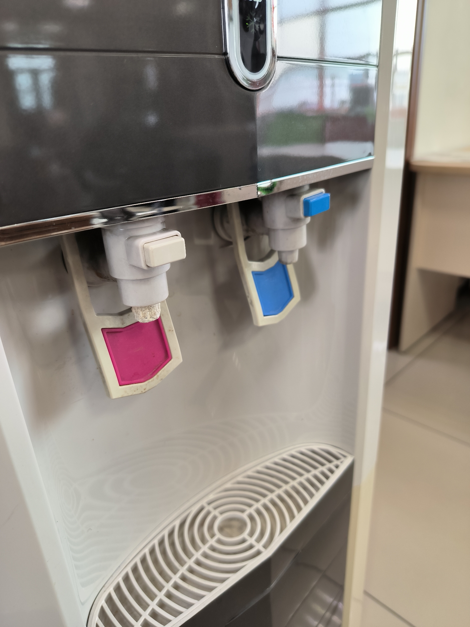 A close-up, eye-level shot shows the dispensing spigots of a white and gray water cooler. Two spigots are visible: one on the left with a pink accent and a white lever, and another on the right with a blue accent and a blue lever. The spigots are made of white plastic. Below the spigots, a white drip tray with a circular grate is present. The background is blurred, showing a hint of an office environment with a desk and possibly a doorway. The image is well-lit, with reflections visible on the glossy surfaces of the water cooler. There are no people or animals in the frame. The scene does not provide any specific clues that would allow it to be located in Yuzhno-Sakhalinsk, Russia, or any other specific city.