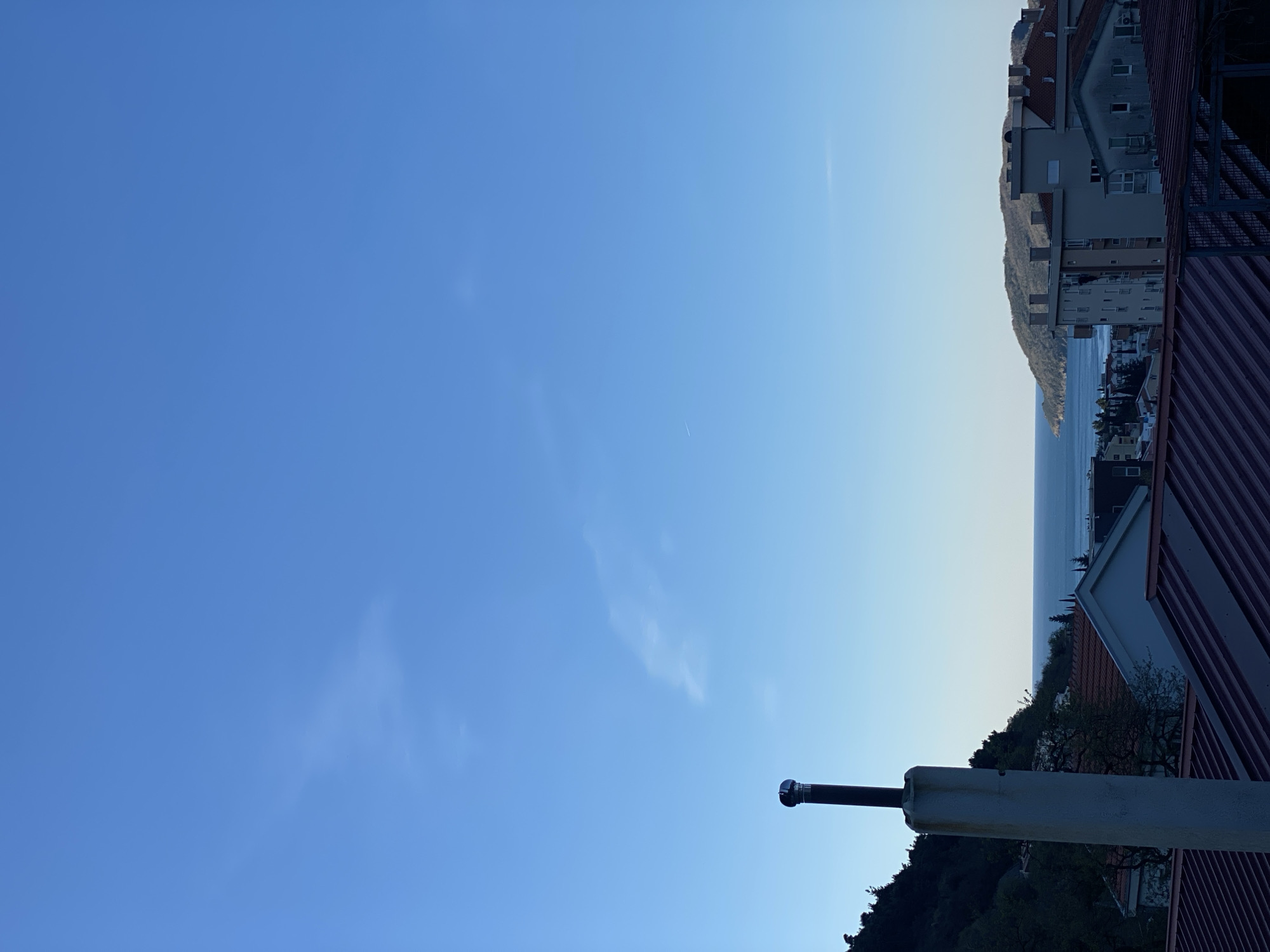 The image shows a view from a rooftop in Budva, Montenegro. A section of a building with reddish-brown corrugated roofing is visible in the lower right. Other buildings are visible in the distance, along the coast. The horizon is a hazy blend of sky and water. A metal pole extends out from the rooftop. The sky is clear blue with wispy clouds.