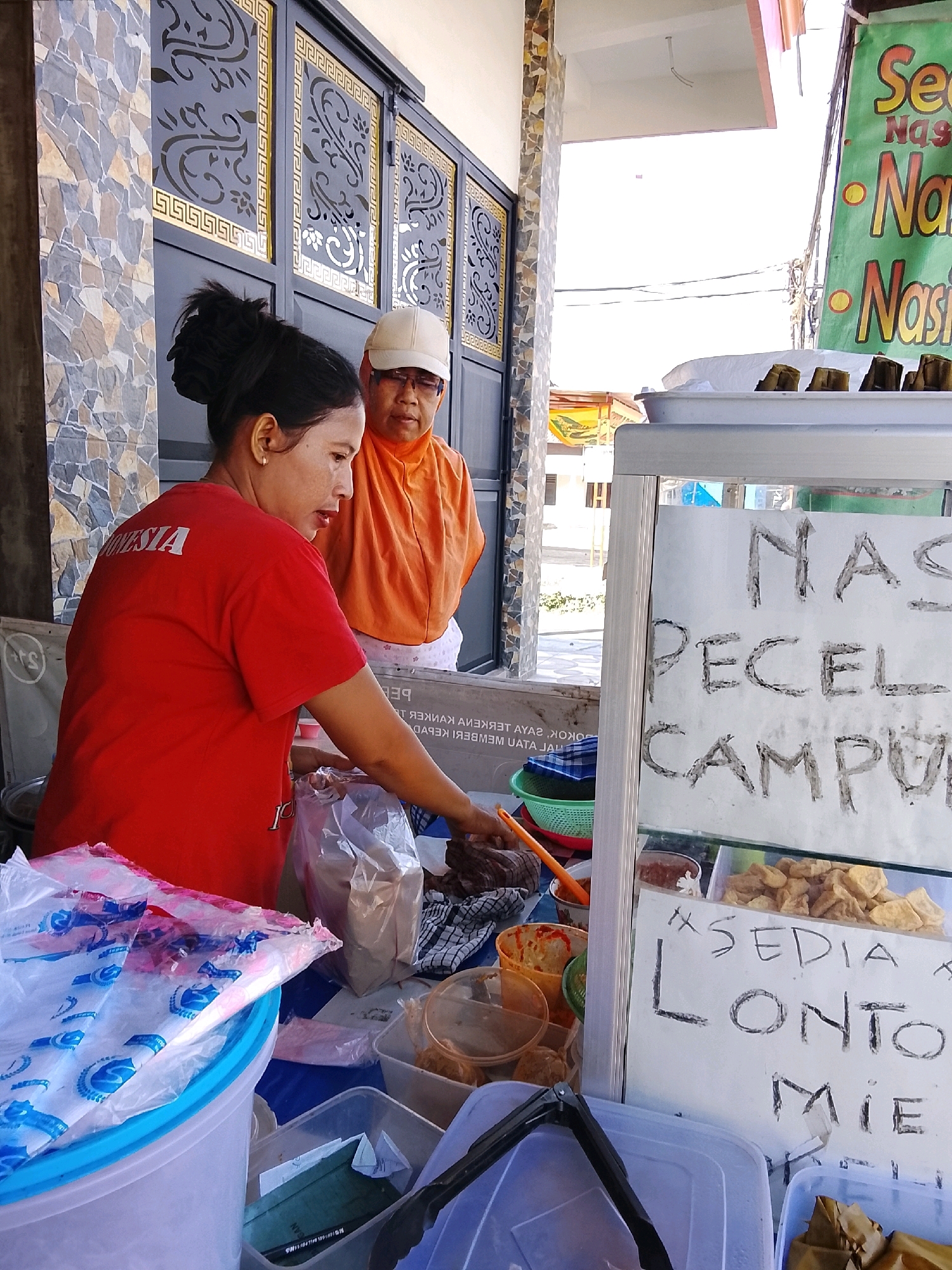 In Dukuhtengah, Indonesia, two women are seen in front of a food stall. The woman in the foreground wears a red t-shirt with "INDONESIA" printed on the back. She is looking down and appears to be arranging items in plastic bags on a counter. Behind her, another woman, wearing an orange headscarf and a white cap, observes. The stall displays various food items, including what appears to be rice and vegetables. A sign on the stall has handwritten text that reads "NASI PECEL CAMPUR" and "#SEDIA LONTONG MIE". The background shows a building with decorative metal gates and patterned stone walls. The scene suggests a busy street food vendor preparing for or serving customers.