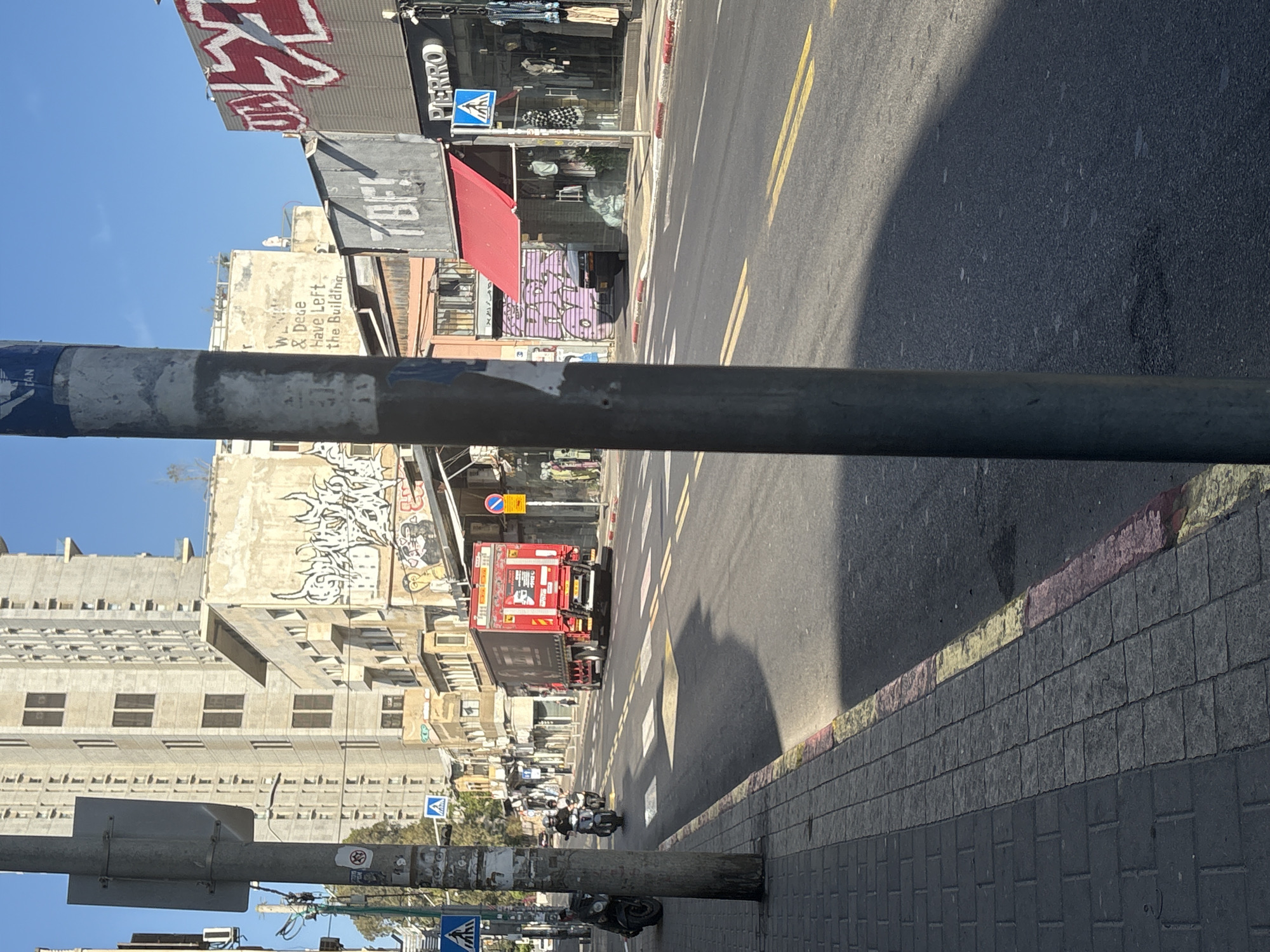 The image depicts a street scene in Tel Aviv, Israel. It is shot from a ground-level perspective, looking along a city street. The foreground is dominated by a pole and sidewalk. The street includes yellow lane markers, and adjacent buildings. A red truck is partially visible in the street. Scooters and people are seen further down the street. Buildings are visible in the background, many of which are covered in graffiti. Pedestrian crossing signs are visible. The sky is blue with no clouds.