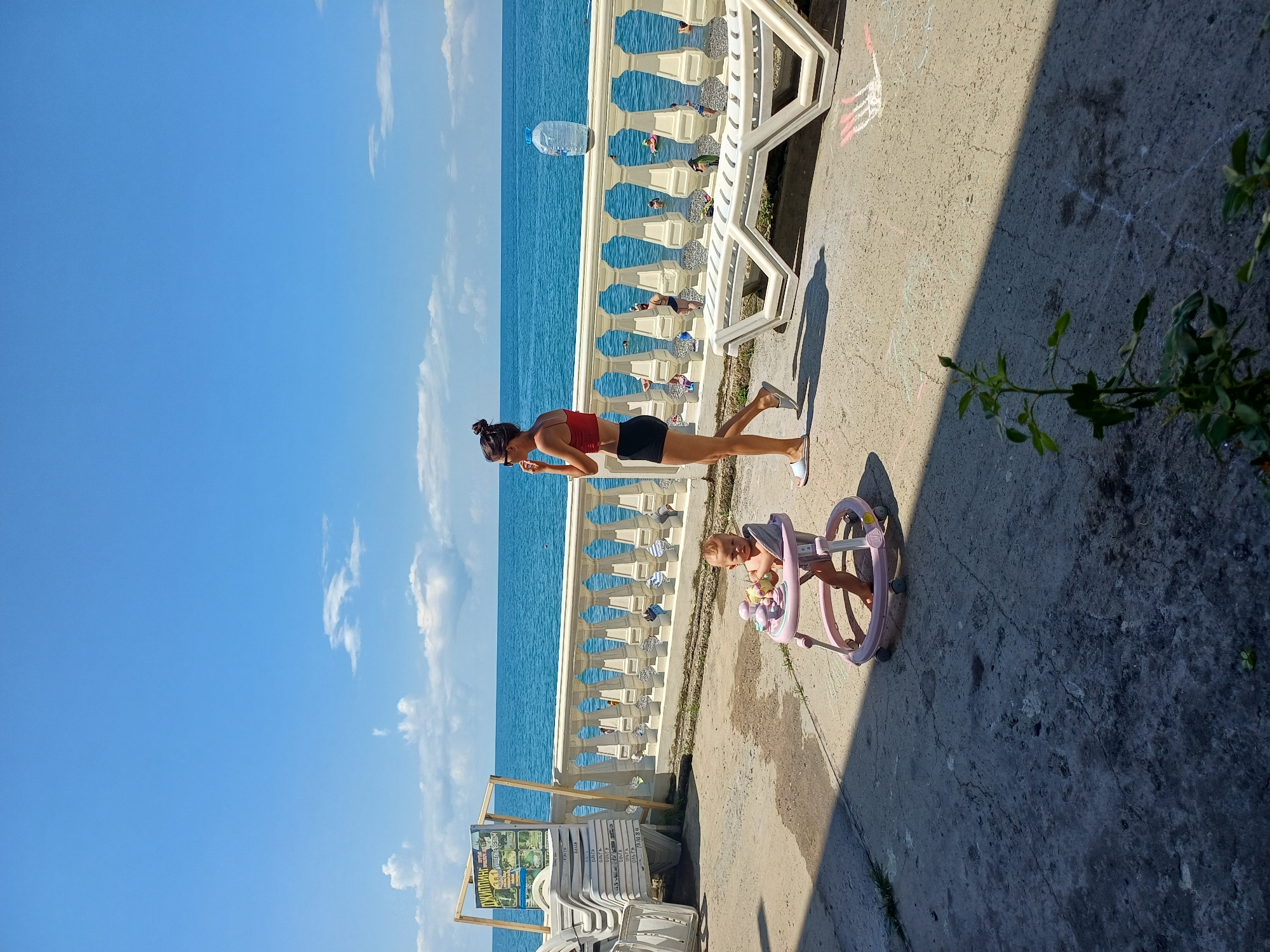 In Sochi, Russia, a woman in a red top and black shorts walks along a sun-drenched promenade next to the Black Sea. She is on the right side of the image, facing left, and appears to be talking on her phone. In the foreground, to the left, a baby sits in a pink baby walker on a concrete surface. The sea is a vibrant blue, with a few white clouds in the sky. Beach chairs are stacked to the left of the image, and a railing separates the promenade from the pebbled beach and the sea. Several people are visible in the water. A large plastic water bottle sits on the railing.