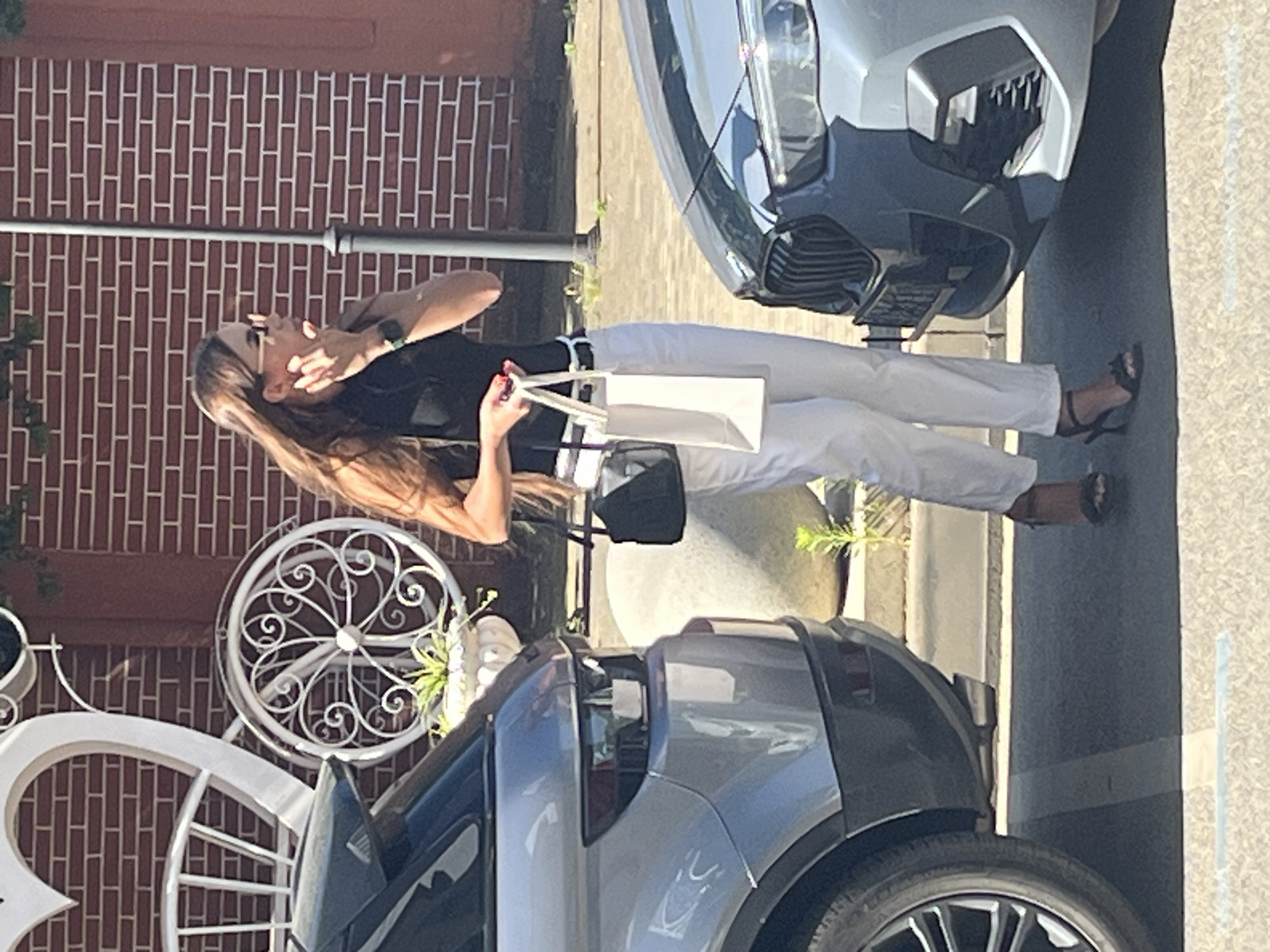A woman is standing outdoors, between two cars. She is wearing a black sleeveless top, white pants, and black high-heeled sandals. She has long brown hair and is wearing sunglasses. She is holding two bags, one black and one white. The scene appears to be in Krasnodar, Russia. The background consists of a brick wall and some decorative metalwork. The environment is sunny.