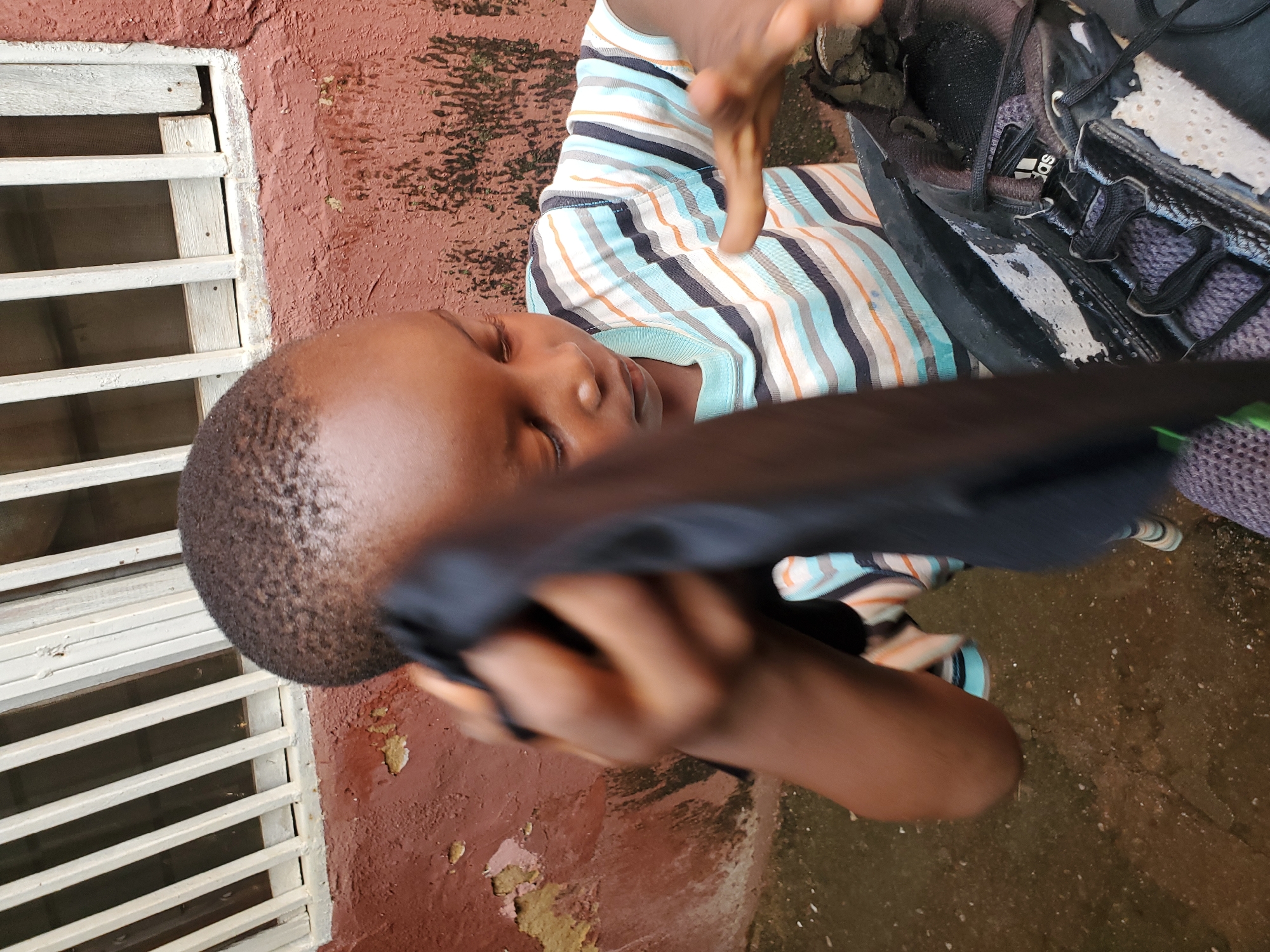 A young boy with short hair and a striped t-shirt is cleaning a black athletic shoe. He is holding a dark cloth in his right hand, using it to wipe down the shoe. The shoe appears to be made of mesh and leather with white accents and black laces. He is focused on the task, with his head bowed. The background is an outdoor scene with a textured, reddish-brown wall and a white barred window frame. The ground appears to be wet and uneven. This scene is likely taking place in an urban area, given the proximity of the wall and window, and the nature of the activity suggests a common domestic setting. The presence of a barred window and the reddish-brown wall are characteristic of buildings found in many parts of Jos, Nigeria, suggesting this photograph was taken there.