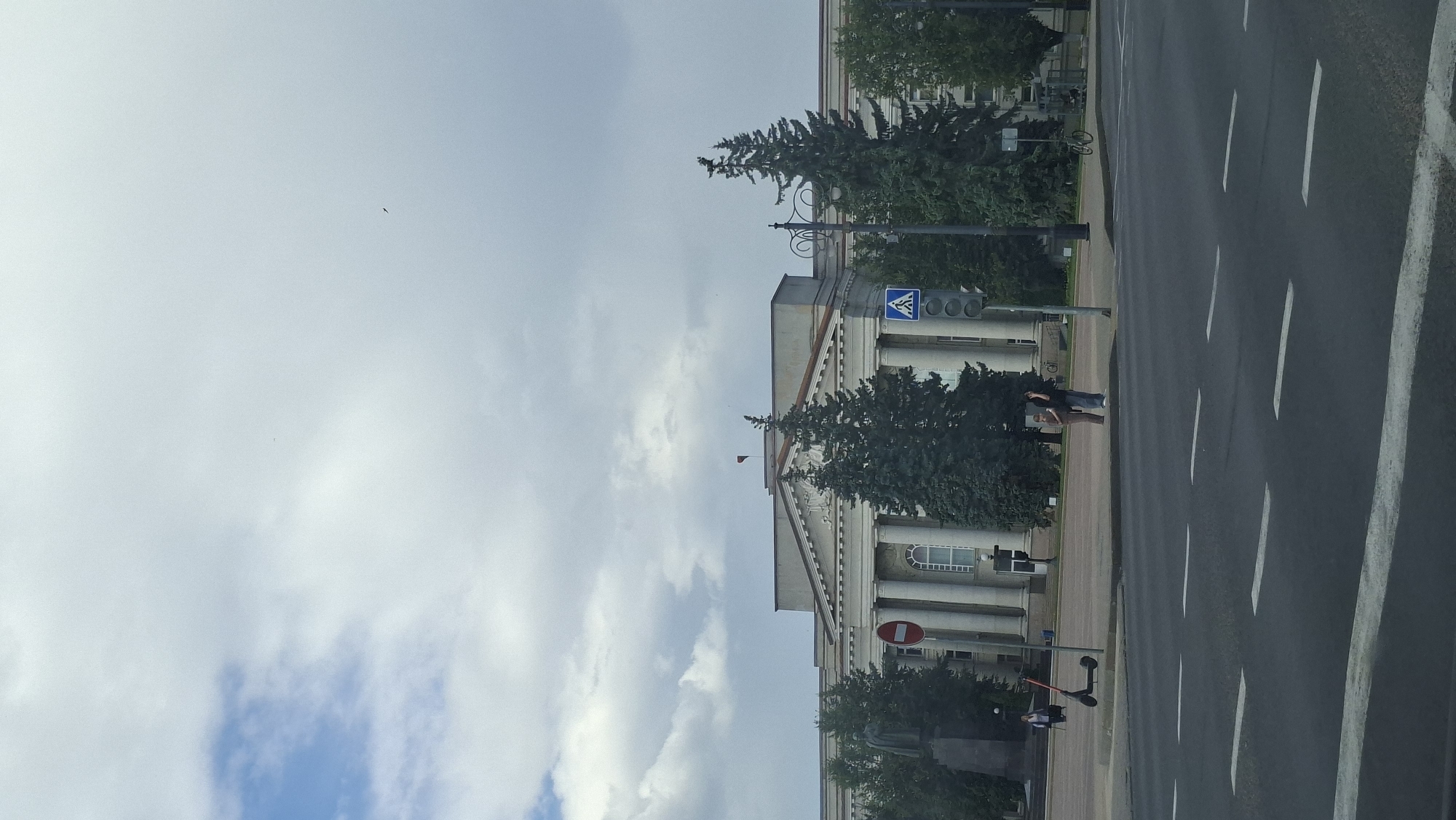 This is a medium shot, taken from a street-level perspective, of a large, neoclassical-style building in Maladzyechna, Belarus. The building features a prominent colonnade and a central triangular pediment. In front of the building, on the left side, is a statue on a pedestal. A red circular sign with a white horizontal bar is visible to the left of the building's entrance. A pedestrian crossing signal with a walking person icon is mounted on a pole in the middle ground, to the right of the building's entrance.
The street in the foreground is paved and marked with white lane lines. A black electric scooter is parked on the sidewalk in front of the building, to the left of the pedestrian crossing. Two individuals stand near the pedestrian crossing, one facing the camera and the other with their back to the camera, both appearing to be waiting. Behind them, evergreen trees are planted along the edge of the sidewalk. To the far right of the image, a lamppost with a curved arm is visible. A bicycle is parked near the lamppost.
The sky is overcast with large, white clouds interspersed with patches of blue. A small, dark object, possibly a bird or an insect, is visible in the sky in the upper right quadrant of the image.