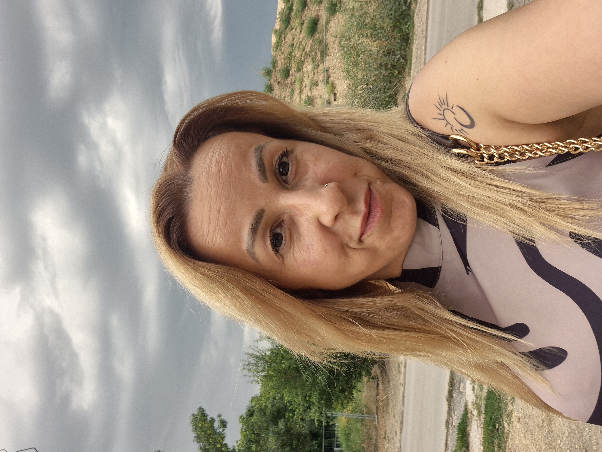 In a close-up, self-portrait style shot, a woman with blonde hair and fair skin is the central subject. She has a small nose ring and a subtle smile. Her hair is worn down, framing her face. She is wearing a top with a light background and dark, abstract patterns. A gold chain is visible over her shoulder. On her upper left arm, she has a tattoo of a sun with rays and a crescent moon shape below it. The background is a mix of greenery, a dirt embankment, and a cloudy sky. The setting appears to be outdoors, possibly in a semi-urban or rural area.