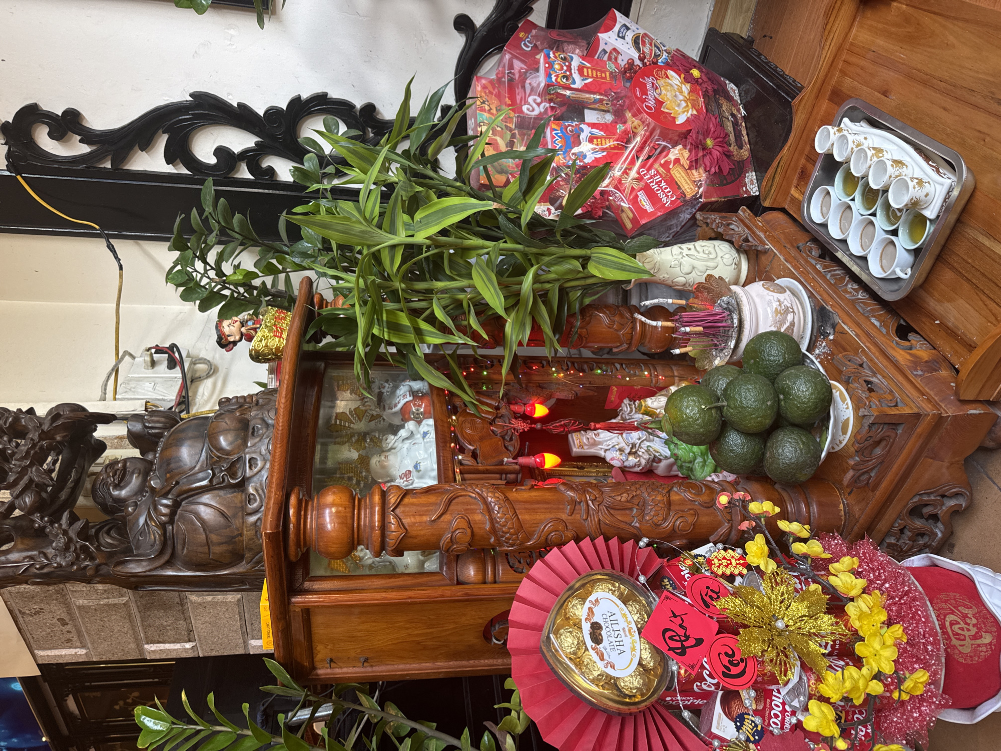 This media file captures a top-down, angled view of a festive display, likely an altar or ceremonial table, adorned with offerings and decorations. The scene is rich with cultural elements, strongly suggesting a celebration such as Tết (Vietnamese Lunar New Year).
The primary subjects include elaborately carved wooden furniture, possibly an altar or cabinet, featuring intricate details like dragons and a dark wooden Buddha-like figure. Lush green leafy plants, resembling lucky bamboo, are prominently placed. Various offerings and decorations fill the space: a large clear plastic bag containing assorted snacks and candies, a tray with several small cups (some filled with liquid, possibly tea), and a plate of green citrus fruits. Burning incense sticks are visible in a decorative vase. Festive red decorations, including a fan-shaped piece, red envelopes (lì xì) with gold script, and artificial yellow flowers with glittery accents, are scattered throughout. A heart-shaped box of "AILISHA CHOCOLATE" and small red string lights contribute to the celebratory atmosphere. Visible through a glass panel are white ceramic figurines.
Visible text includes "AILISHA CHOCOLATE," "ASSORTED COOKIES," "TRANG LONG," "ORIO," and "noco ChocoPle Ple," along with Vietnamese characters on the red envelopes, characteristic of New Year greetings. The overall arrangement, color scheme of red and gold, and specific items like lì xì and offerings, point to a traditional festive event, emphasizing prosperity and good fortune. The location context is unknown.