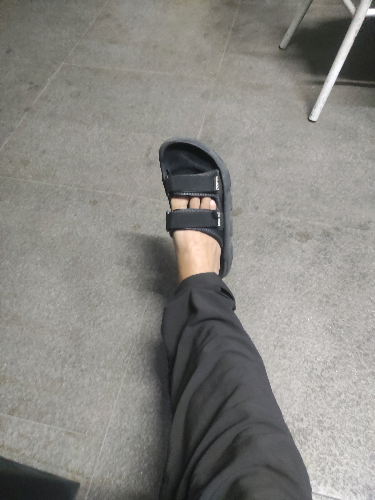 The image shows a close-up, first-person perspective of a person's left foot wearing a black sandal with two adjustable straps. The sandal is placed on a grey, tiled floor with visible grout lines. The person is also wearing dark-colored pants that extend down the leg, partially obscuring the view. In the background, out of focus, are the white legs of a chair. The scene appears to be indoors, possibly in a public space in South Jakarta, Indonesia, given the floor tiling and furniture.