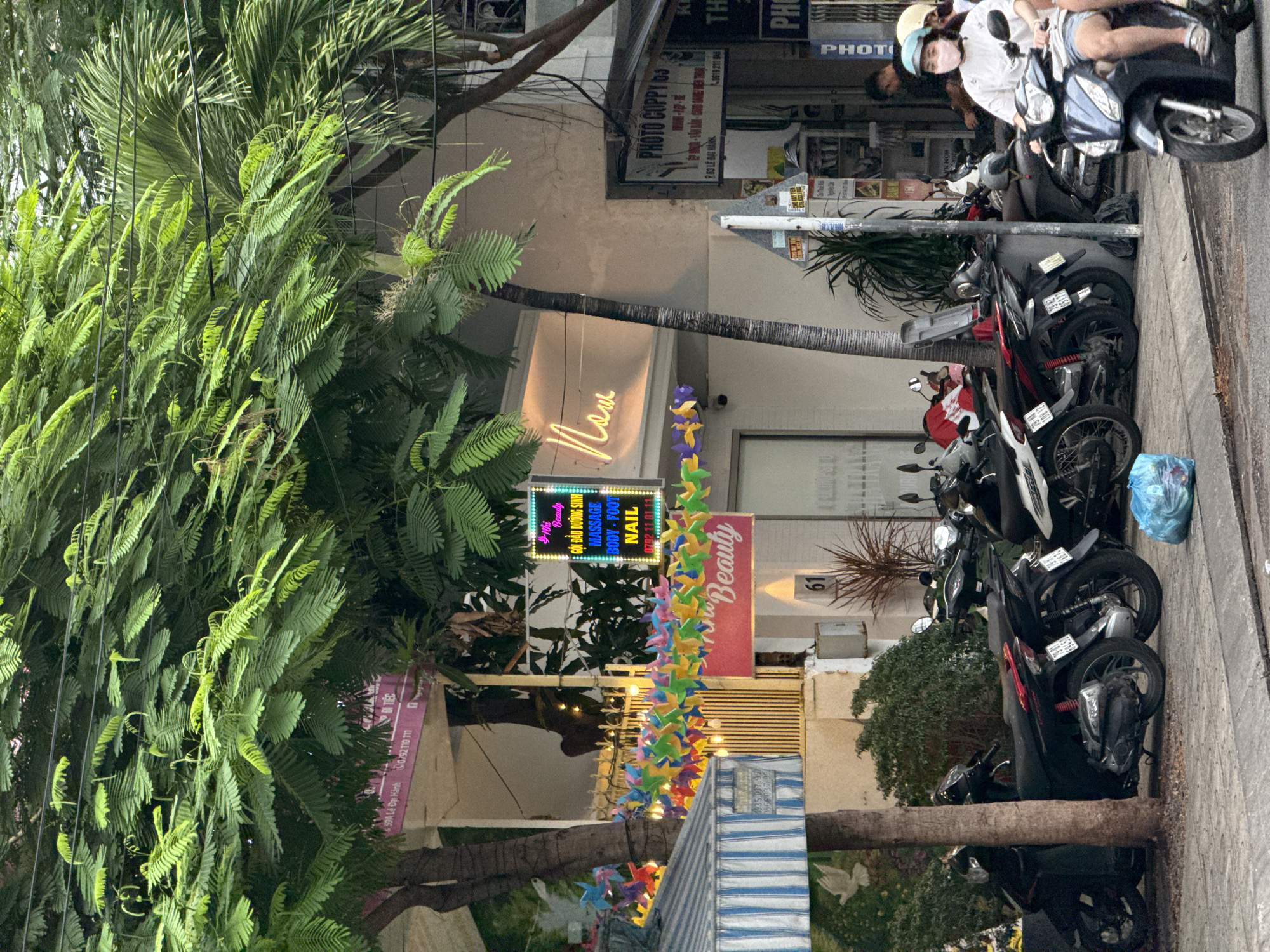 A street scene in Nha Trang, Vietnam, shows a row of parked motorcycles on a sidewalk in front of shops. A bright pink sign above the motorcycles reads "Beauty" with smaller text indicating "MASSAGE BODY - FOOT NAIL". A neon sign also displays "Now". Colorful pinwheels are strung across the scene, adding a festive touch. The shops appear to be open, with awnings and advertisements visible. A person wearing a white shirt, denim shorts, and a light blue helmet rides a motorcycle away from the camera. The street is paved, and there are trees and power lines overhead.