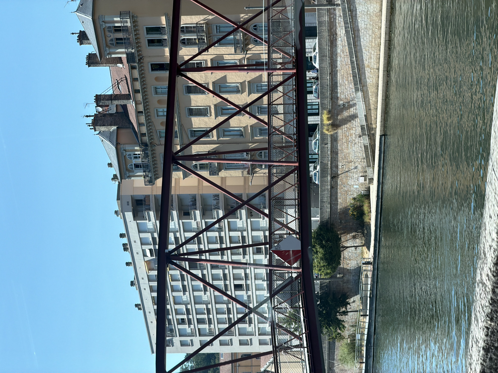 The image captures a view of the Rhône River in Lyon, France, seen from a boat. A modern, red metal truss bridge crosses the river in the foreground, spanning across the water and the riverside embankment. Behind the bridge, a multi-story, light-colored apartment building with numerous balconies and uniformly spaced windows dominates the background. To the right of the apartment building are older, ochre-colored buildings with ornate architectural details, including balconies and classical-style window surrounds. The sky is a clear, bright blue. The river's surface is calm, reflecting the sky and the surrounding structures. The embankment features a paved walkway with railings and some trees and greenery. Cars are visible parked on a road above the embankment, behind the bridge. A small, triangular red and white sign is affixed to the side of the bridge.