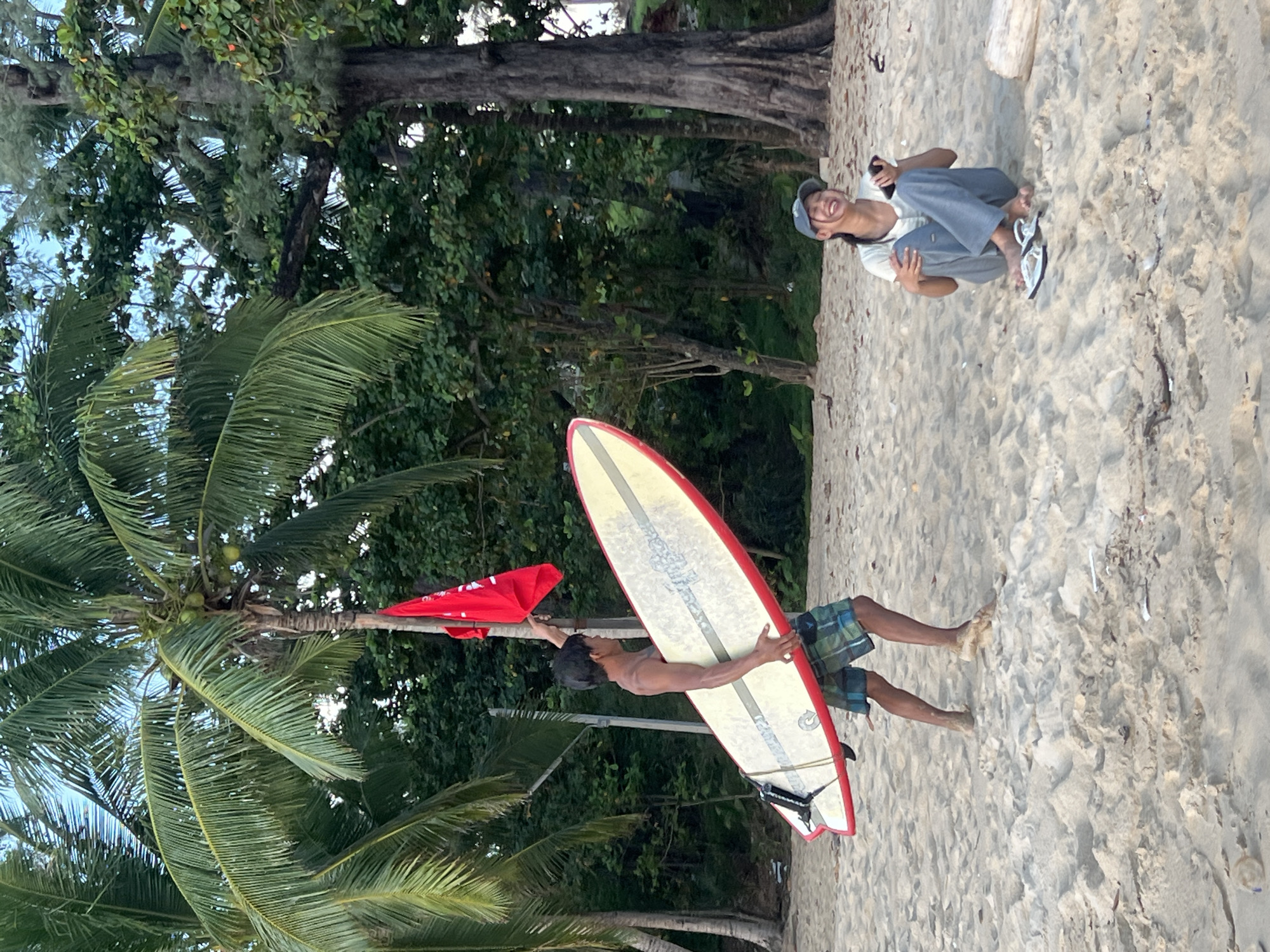 In the city of Choeng Thale, Thailand, on a sandy beach, a young man walks carrying a surfboard and a red flag on a pole. He is wearing checkered shorts. To his right, a young woman sits on the sand wearing a hat, t-shirt, and pants, holding a phone. She appears to be smiling and laughing. The background consists of lush green palm trees and dense foliage, with the ocean not visible. The lighting suggests it is either early morning or late afternoon.