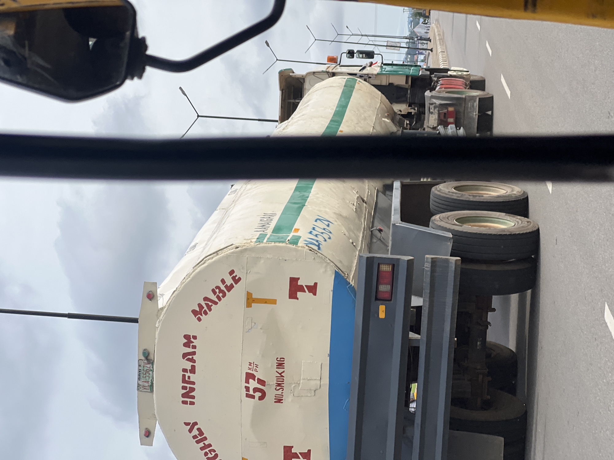The image depicts a tanker truck with the words "HIGHLY INFLAMMABLE" written on its side, suggesting it is transporting hazardous materials. The truck is seen on a road, likely in Jalingo, Nigeria, indicated by the license plate with "Yola" and "AJG" which are associated with Adamawa state. The license plate also shows "YCA-5567V". On the side of the tanker, there is a speed limit indicator of "57 KM/PH" and a "NO SMOKING" sign. There are also red "T" symbols on the tanker, which are warning symbols for flammable materials. The environment appears to be an urban or semi-urban area with streetlights visible in the background and other vehicles on the road, including a yellow vehicle in the foreground that is partially obstructing the view. The sky is cloudy. The image is taken from inside another vehicle, with a black bar and a yellow object suggesting a window or interior of the vehicle.