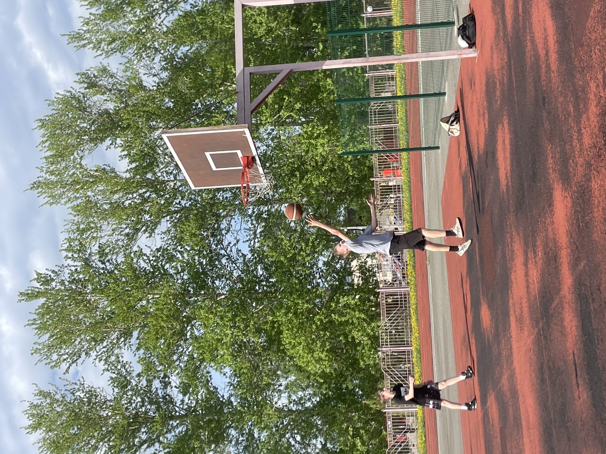 The image depicts a basketball court in Zav'yalovo, Russia. A young man is in mid-air, reaching to shoot a basketball towards the hoop. Another person stands on the court nearby. The court is red with some darker shaded areas and bordered by a green metal fence. Trees with green foliage are visible in the background. A few bags are on the ground near the fence. The sky is partly cloudy.