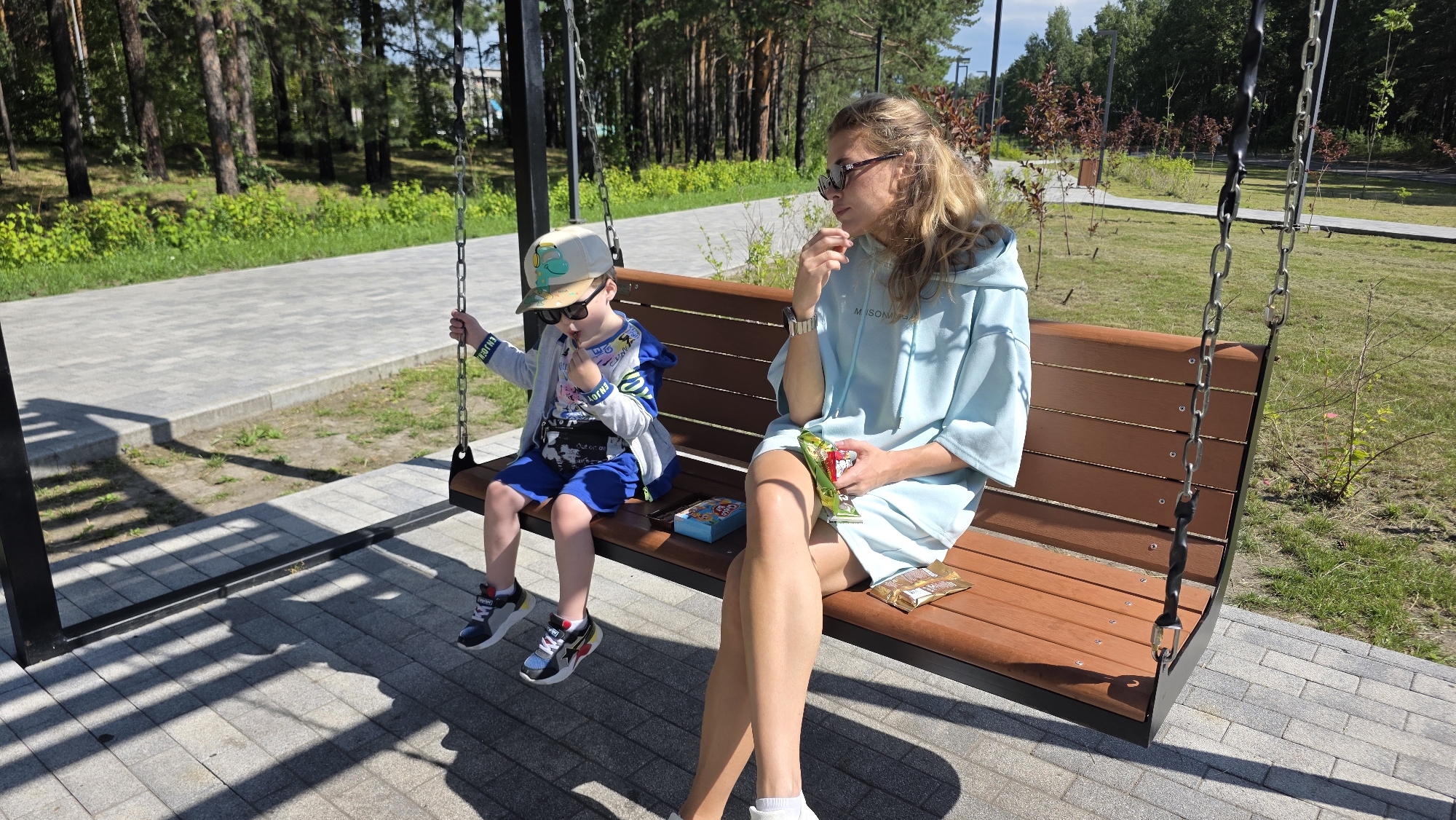 A woman and a child are sitting on a wooden swing in a park. The woman is wearing a light blue hoodie and sunglasses, and she is holding a snack. The child is wearing a hat, sunglasses, a gray jacket, and blue shorts, and he is also holding a snack. There is a box of snacks on the swing between them. The park has paved walkways and trees in the background. The scene appears to be in the city of Angarsk, Russia.
