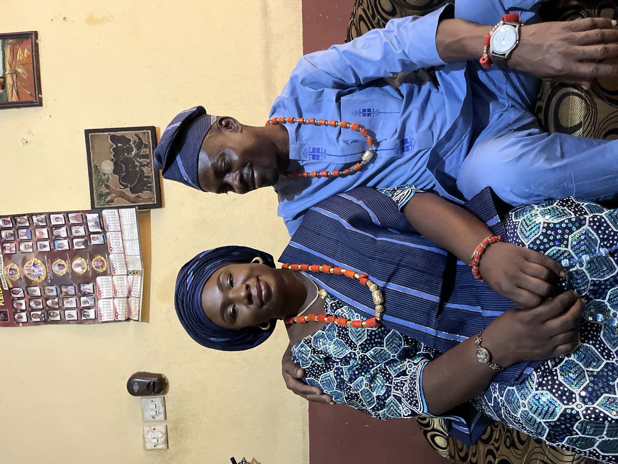 A photo shows a man and a woman indoors. The man is wearing a light blue shirt and a matching hat with blue stripes, and an orange beaded necklace. He has a wristwatch with a red band on his left wrist. The woman is wearing a dark blue top with blue stripes and a matching head wrap. Her top is patterned with white dots and blue embellishments. She also wears an orange beaded necklace and a bracelet on her left wrist. The two individuals are sitting close together. The woman is looking at the camera and smiling. The background is a yellow wall with two framed pictures and a calendar hanging on it. The location is Rafin Bauna, Nigeria, although this information cannot be visually confirmed from the image.