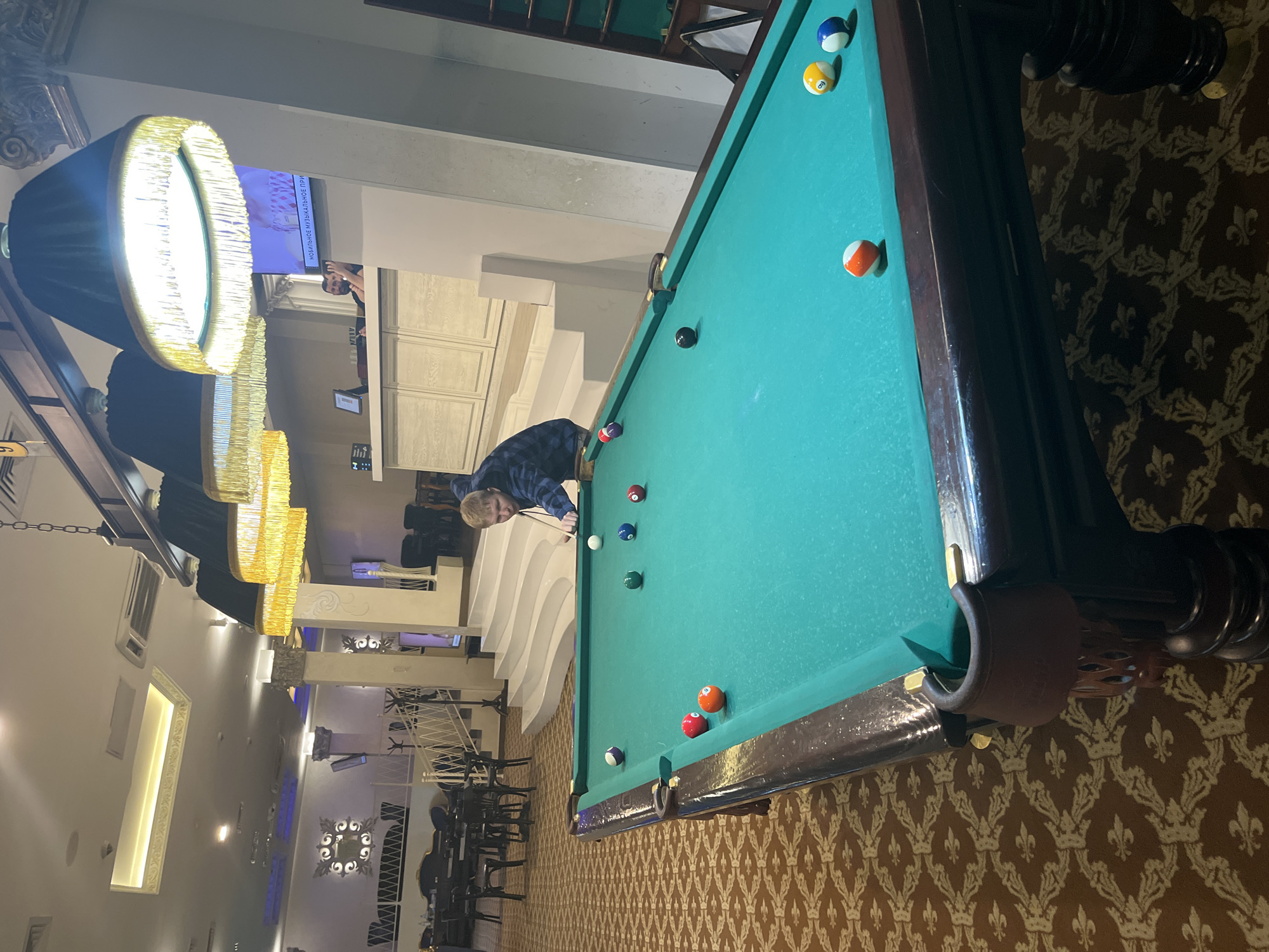 A medium, full shot taken in Sochi, Russia. A man bends over a billiards table, aiming his cue. The table has a green felt surface and several billiard balls scattered across it. The room is decorated with elaborate chandeliers and an ornate, patterned floor. Seating is visible in the background.