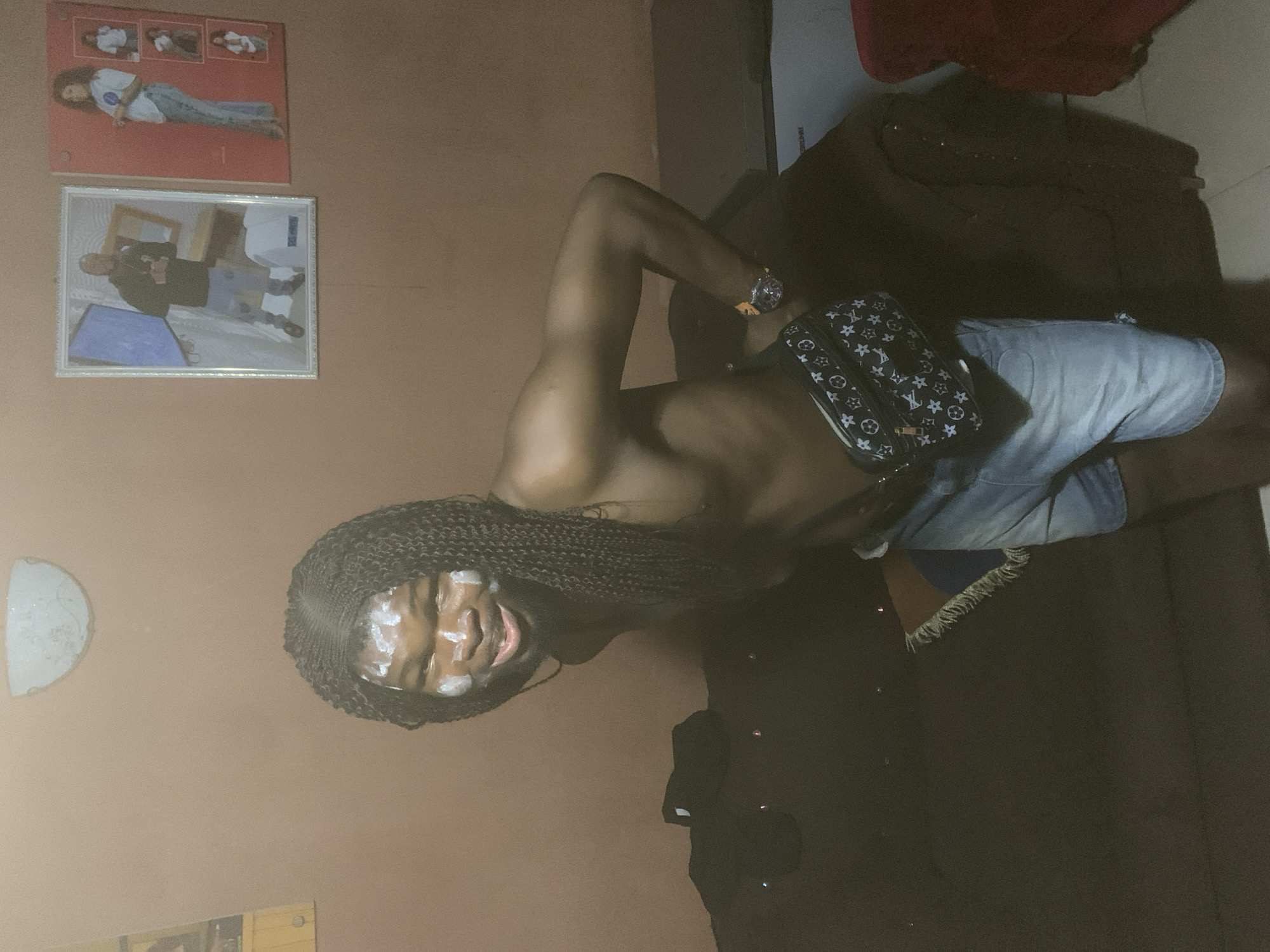 The media file presents an indoor portrait, originally rotated ninety degrees counter-clockwise. The main subject is a person with dark skin and long, dark braided hair, standing shirtless and displaying a muscular physique, particularly in the arms and shoulders. They are wearing light-wash denim shorts and a black fanny pack, adorned with a white repeating pattern resembling a designer monogram, across their chest. A wristwatch is visible on their left wrist. White cream or paste is applied to their forehead and cheeks, and they are looking directly at the camera with a smile or smirk.
The background features a plain, light brown wall. To the subject's left, a white semi-circular fixture or decoration is mounted on the wall. Above the subject's head, two pictures hang on the wall: an upper red board displaying a larger image of a person lying down in a white shirt and jeans, along with three smaller thumbnails; and below it, a white-framed picture showing a person in a dark shirt holding an object, with a blue item in the foreground. To the subject's right, a dark-colored sofa or armchair, possibly covered with a blanket, is visible. The lighting is dim, indicating an indoor setting with low light, perhaps during the evening. No explicit text is discernible within the image, apart from the decorative pattern on the fanny pack. The location context provided is Onitsha, Nigeria.