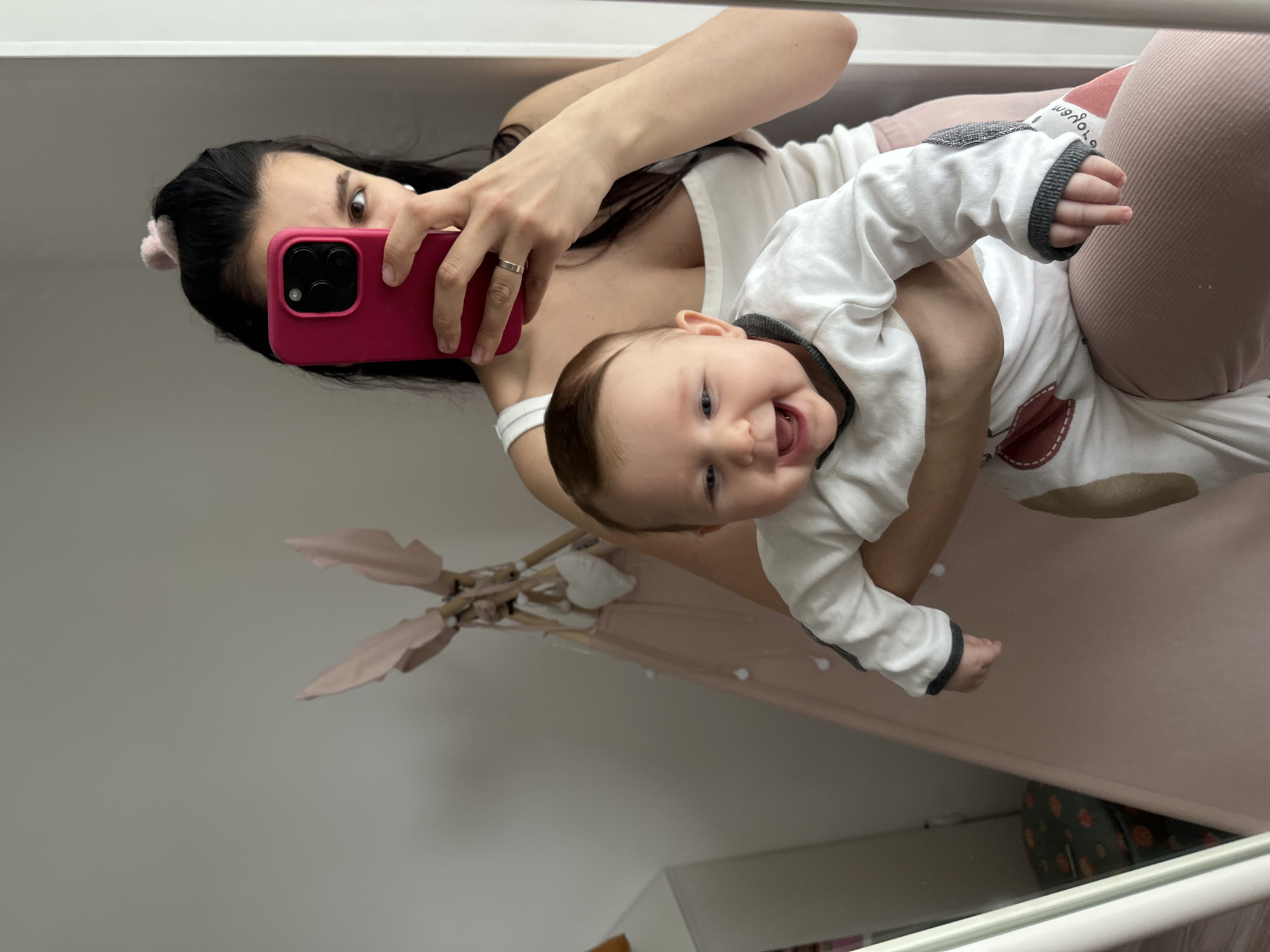 This image, captured in Chaikovsky, Russia, shows a mother holding her baby and taking a mirror selfie in what appears to be a home setting. The image is rotated 90 degrees clockwise.
The central subjects are a woman and an infant. The woman, likely the mother, has long dark hair, and her face is partially visible behind a bright pink smartphone she holds to take the picture. She wears a white top and light pink/peach trousers, with a ring visible on her left hand. The baby, an infant with light brown hair, is held in her arms and is smiling widely with an open mouth, appearing very happy. The baby wears a white long-sleeved onesie with gray trim on the cuffs and collar, and some subtle patterns or patches on the fabric.
The activity taking place is a joyful interaction between mother and child, captured through a mirror reflection. The baby's happiness is a notable detail, creating a warm and affectionate atmosphere.
The setting appears to be indoors, likely a bedroom or nursery, indicated by elements reflected in the mirror. In the background, a light pink play tent or teepee with wooden poles is visible. Below the baby's feet, a patterned sheet or blanket, possibly from a bed or crib, adds a touch of color to the scene. The room walls are a light, neutral color.
There is some indistinct, rotated text on the baby's clothing that is difficult to decipher, possibly "wovojo" or similar. No other clear text is visible. The time of day appears to be daytime, given the general lighting, though no specific light sources are evident.