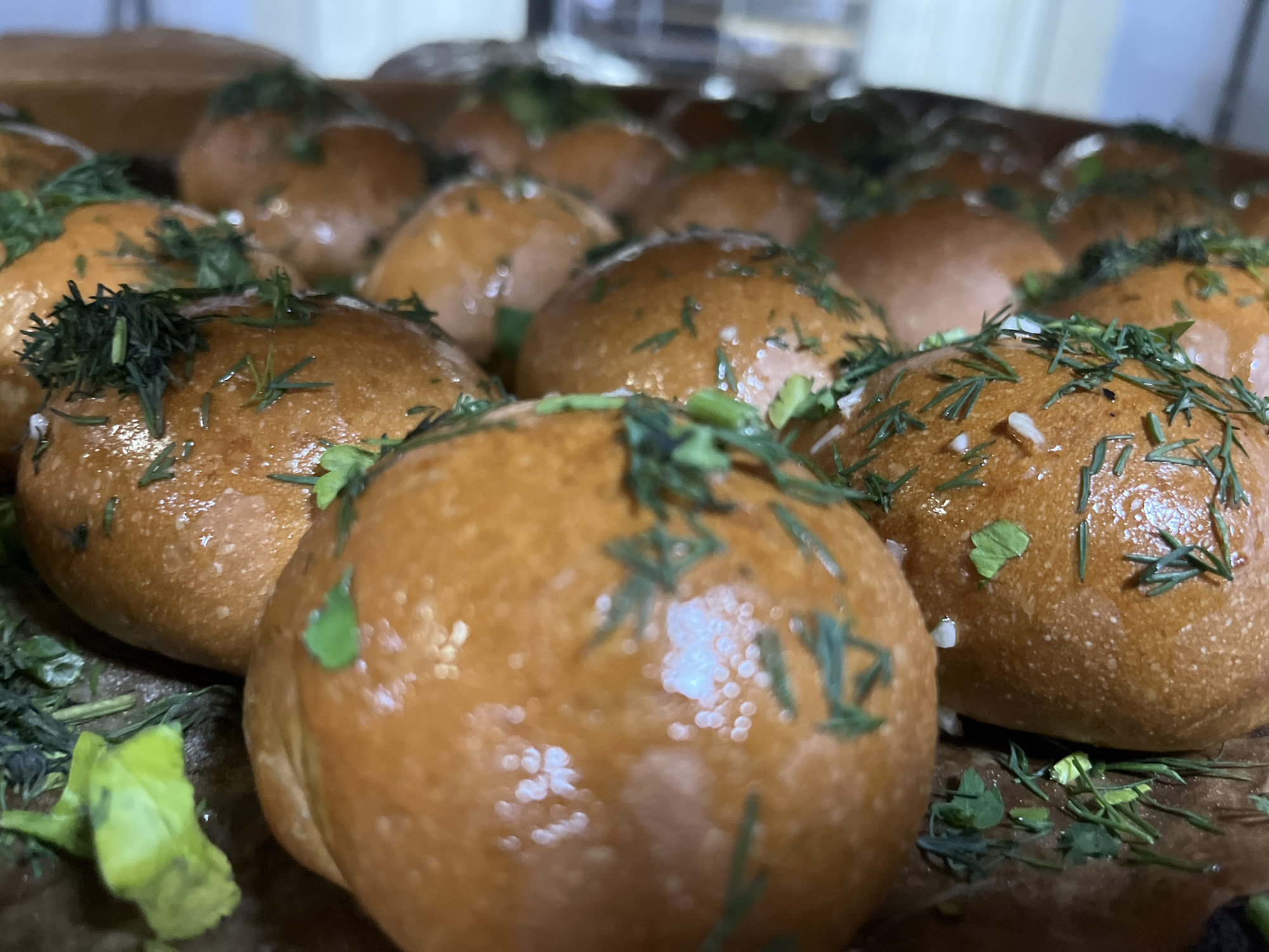 The image depicts a close-up view of several golden-brown buns, likely freshly baked, arranged closely together. They are topped with a scattering of fresh green herbs, possibly dill and parsley. The surface of the buns appears glossy, suggesting they may have been brushed with butter or an egg wash. The photo was taken in Novopokrovskaya, Russia.