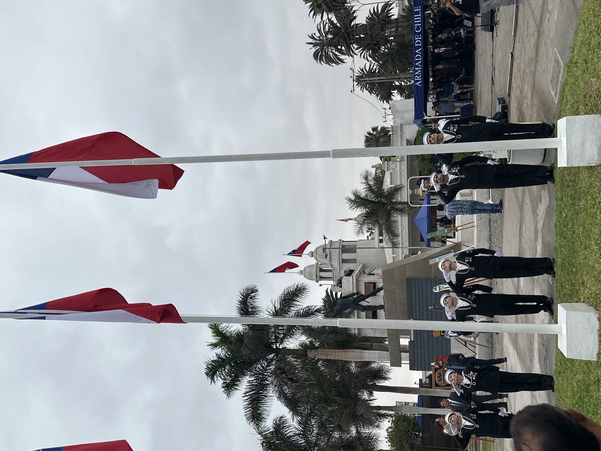 The image depicts a ceremony in Arica, Chile, likely involving the Chilean Navy ("Armada de Chile" is visible on a banner). Several navy personnel in white sailor hats and dark uniforms are saluting. Chilean flags are prominent on flagpoles and atop a white building with domes, possibly a historical or government structure. Palm trees dot the landscape. A bronze statue of a figure in mid-leap is visible in front of the white building. An audience is gathered behind the navy personnel. The sky is overcast.